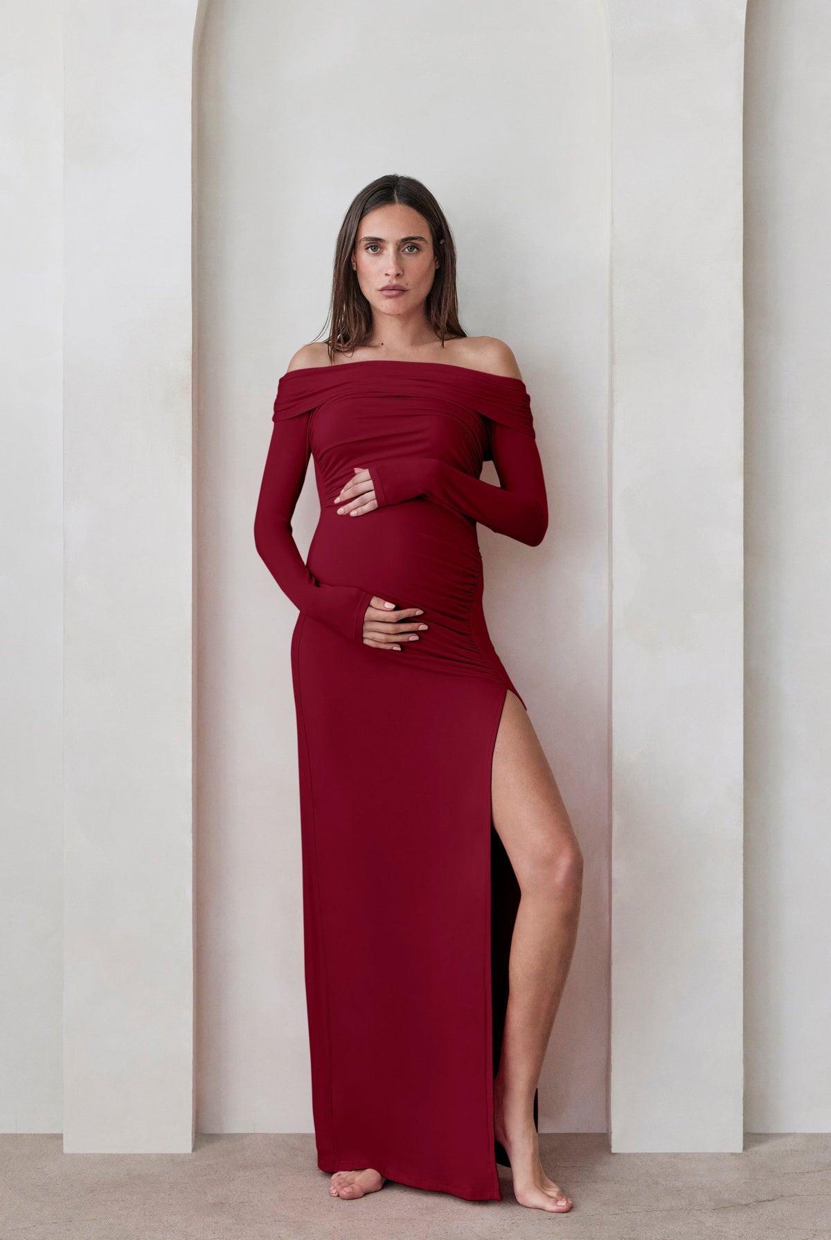 A pregnant woman wearing a Bumpsuit burgundy off the shoulder maxi dress with high leg slit against a beige feature wall