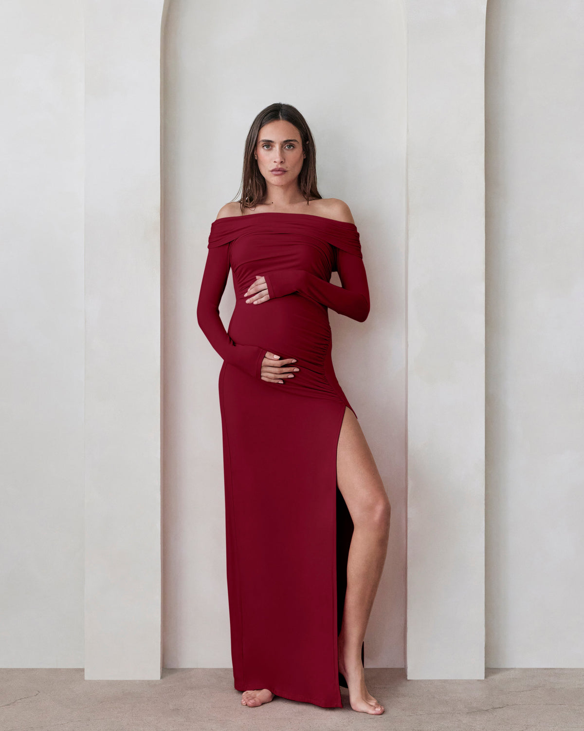 A pregnant woman wearing a Bumpsuit burgundy off the shoulder maxi dress with high leg slit against a beige feature wall