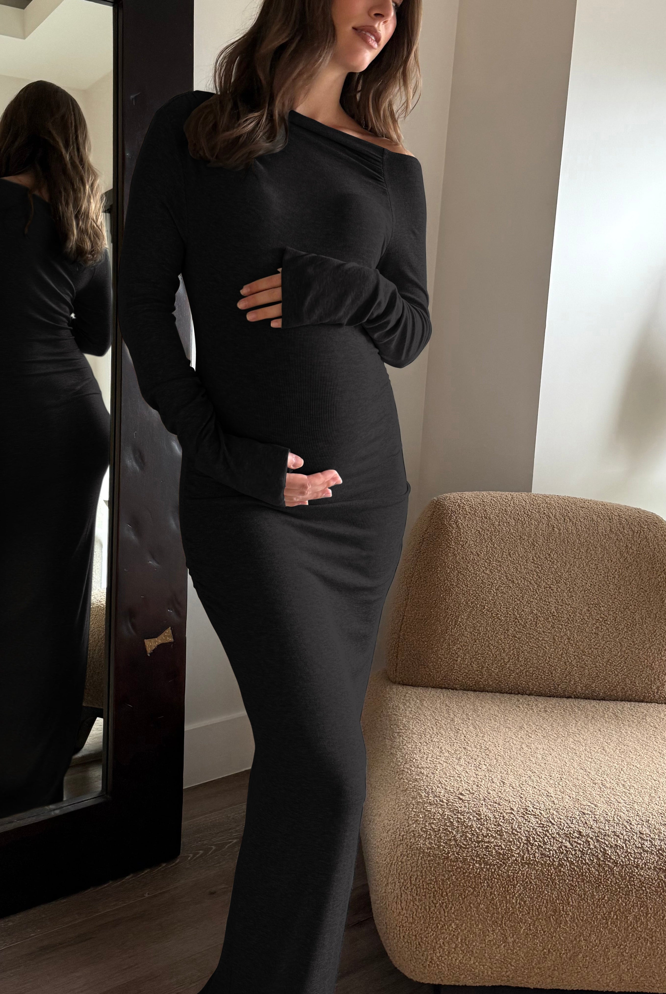 A pregnant woman wearing a black long-sleeve maternity dress stands in a modern room next to a large mirror, showing both her front and back view in the reflection. A beige boucle armchair and a side table with a vase are visible in the background.
