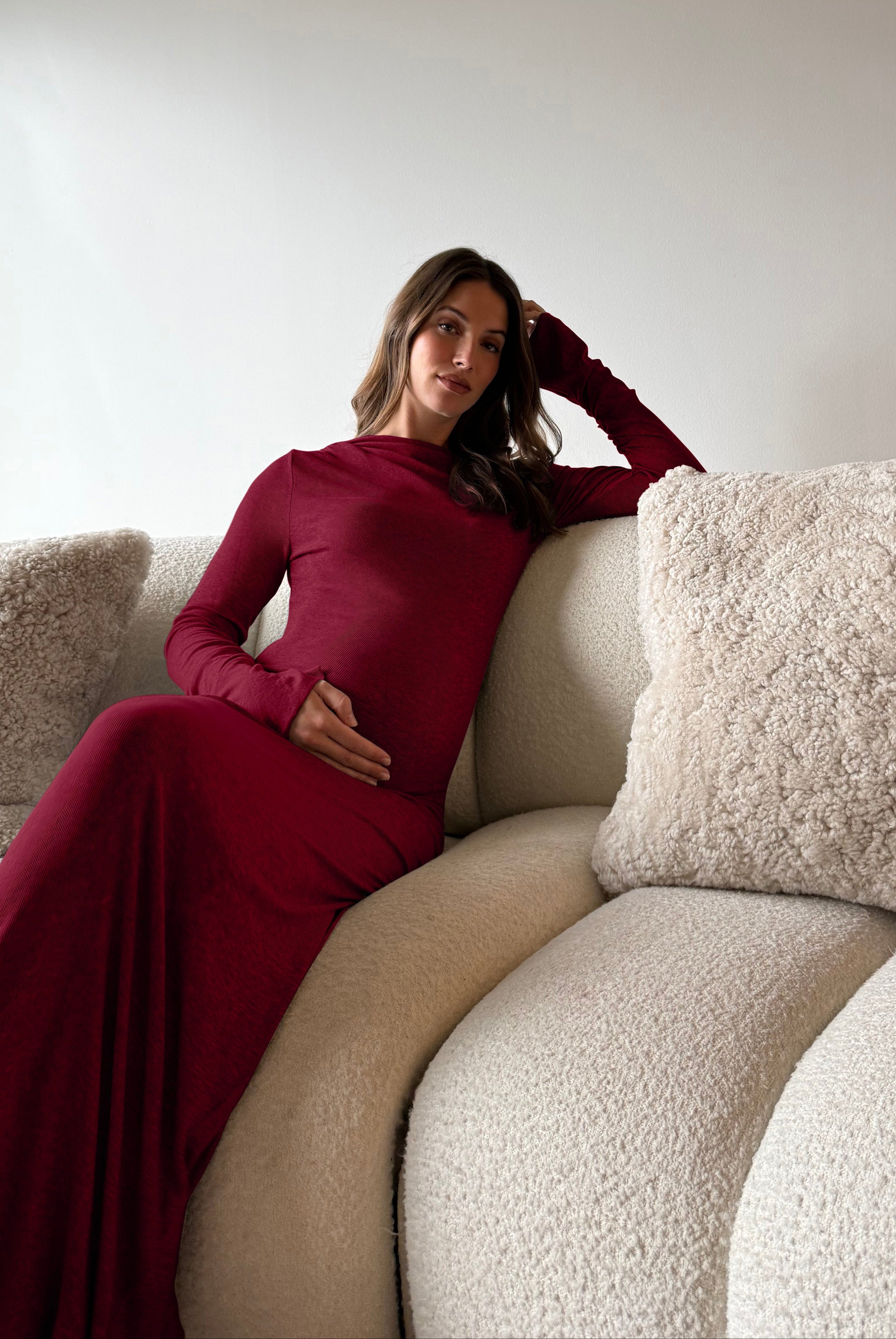 A woman wearing a Bumpsuit long-sleeve soft rib burgundy maxi dress reclines on a cream boucle sofa with textured throw pillows.