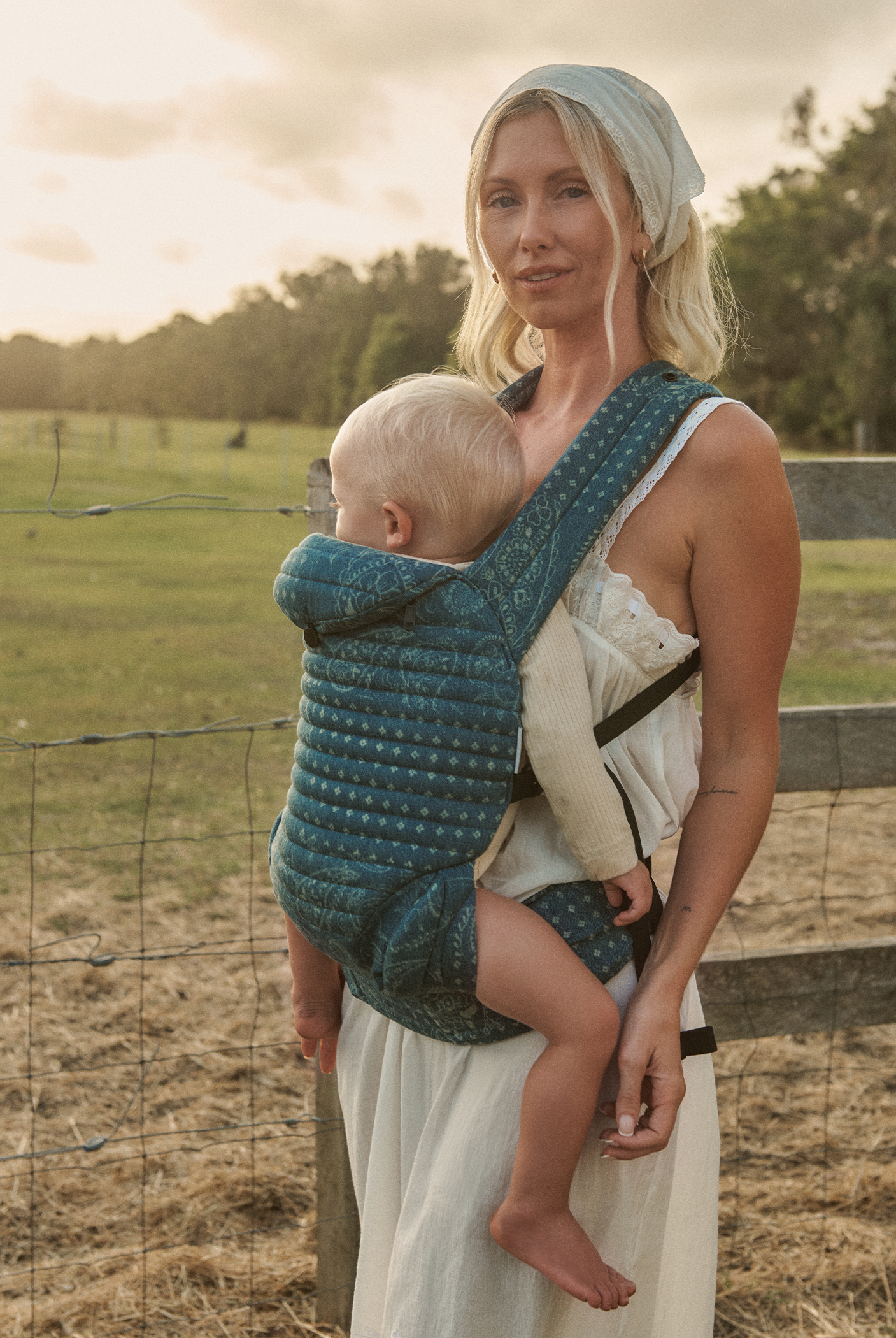 Woman holding a baby in a Bumpsuit armadillo carrier outdoors with a fence and trees in the background