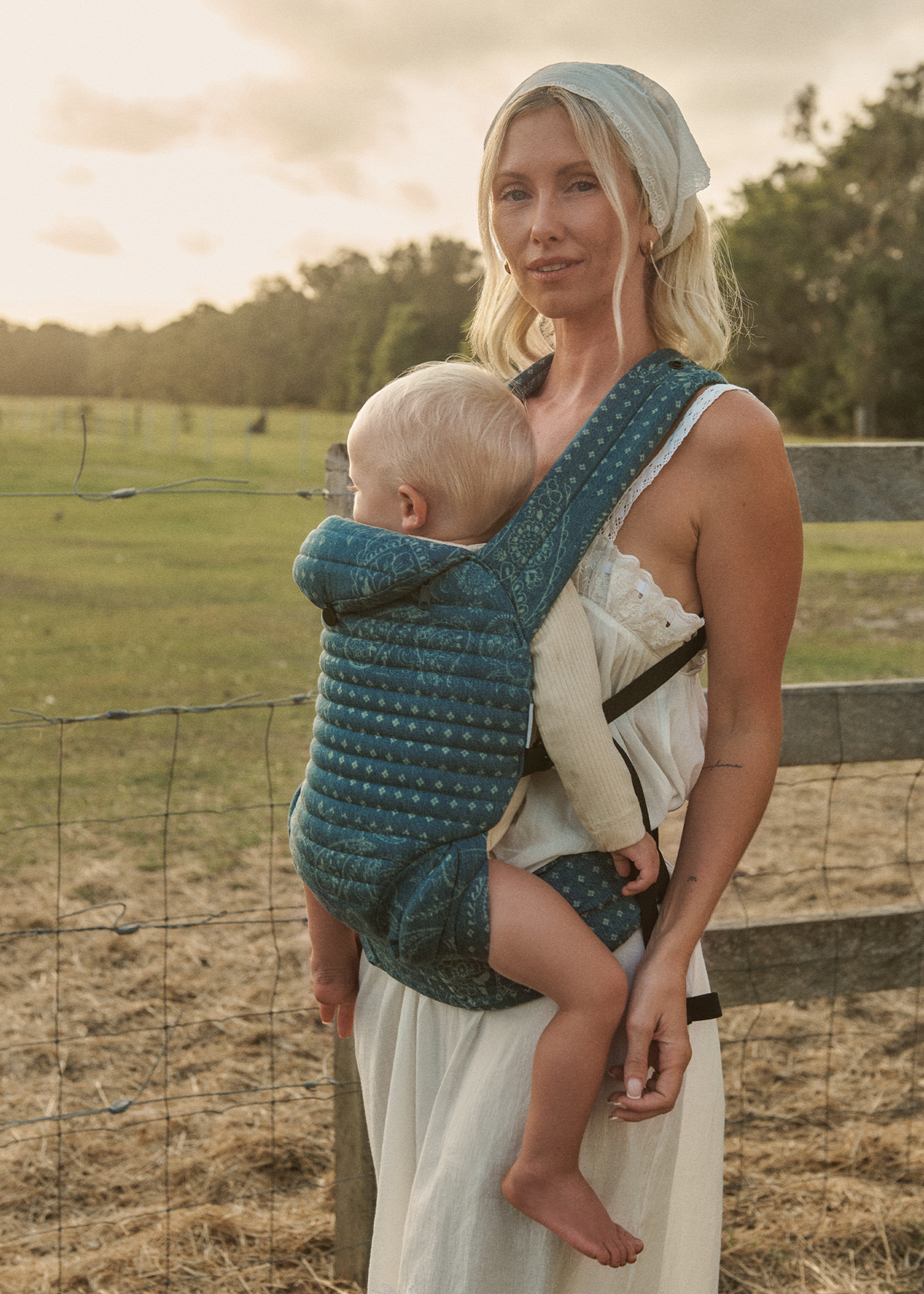 Woman holding a baby in a Bumpsuit armadillo carrier outdoors with a fence and trees in the background