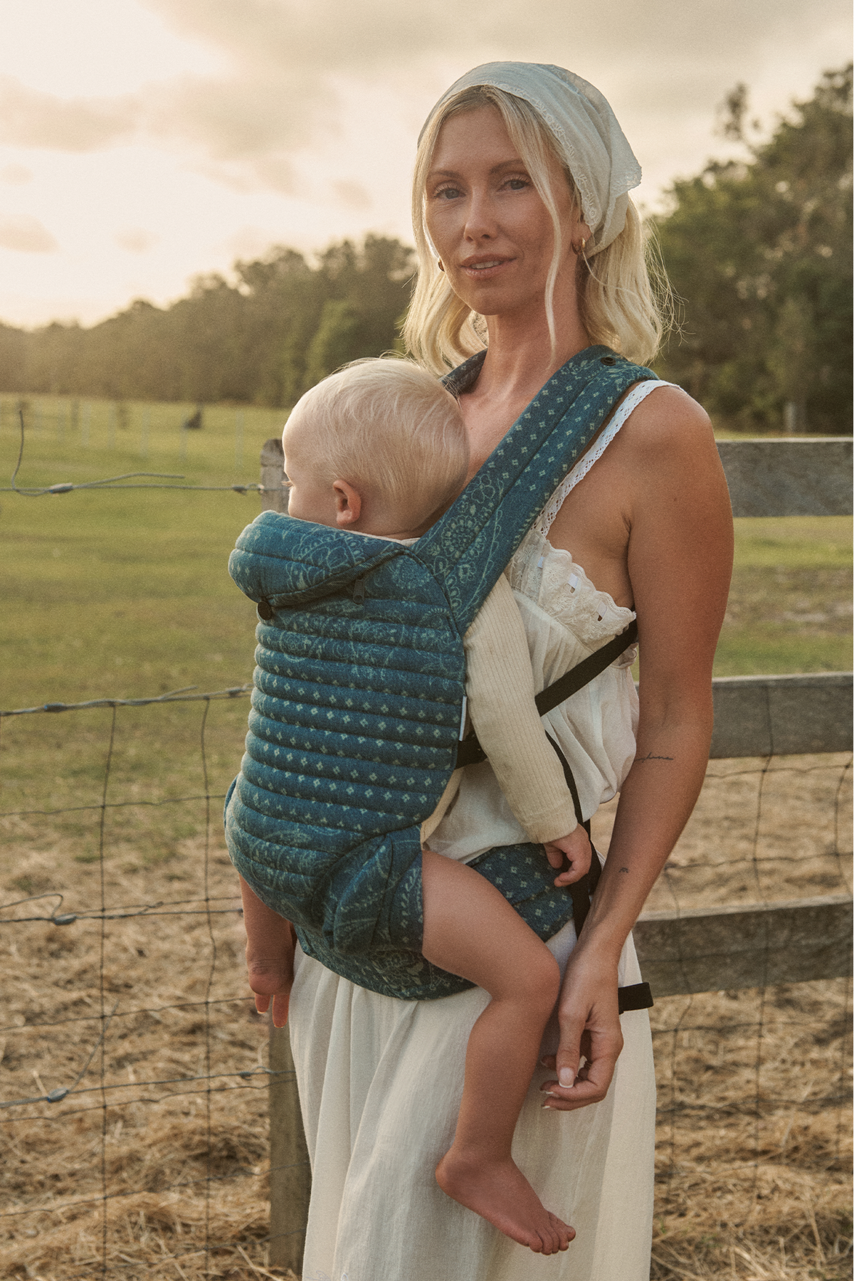 Woman holding a baby in a Bumpsuit armadillo carrier outdoors with a fence and trees in the background
