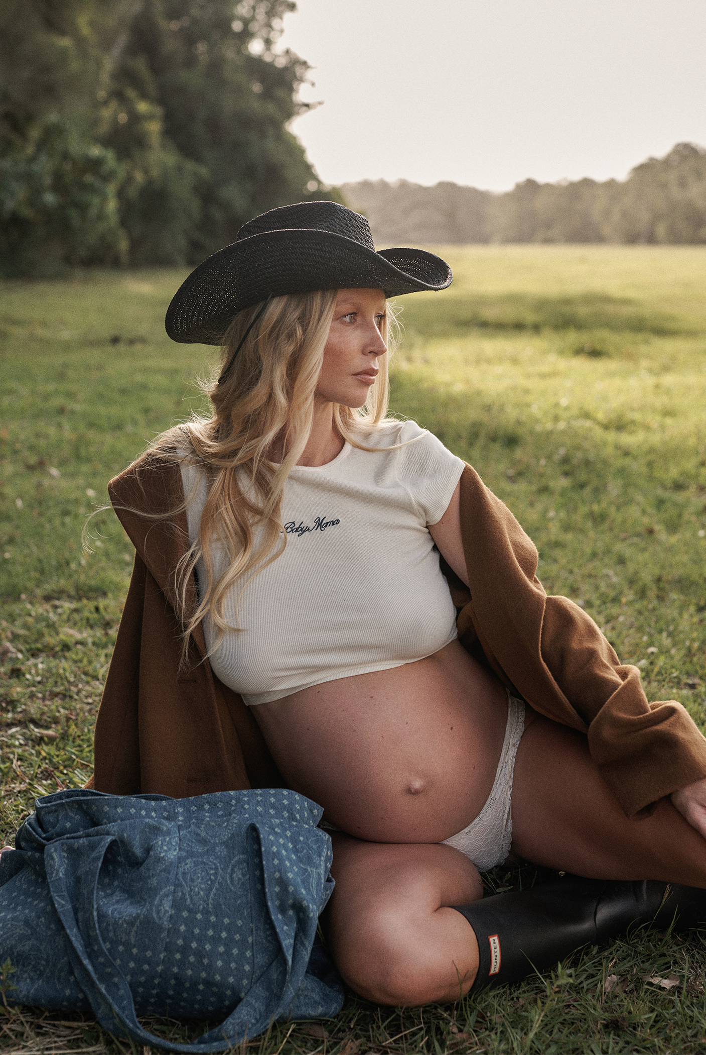 Pregnant woman sitting in a field wearing a wide-brimmed hat and brown coat.