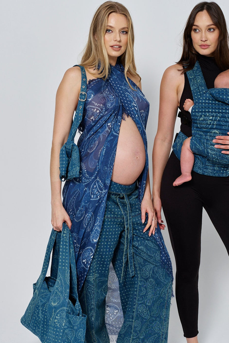 Two women wearing spell print denim outfits with a bumpsuit baby carrier and handbag on a white background