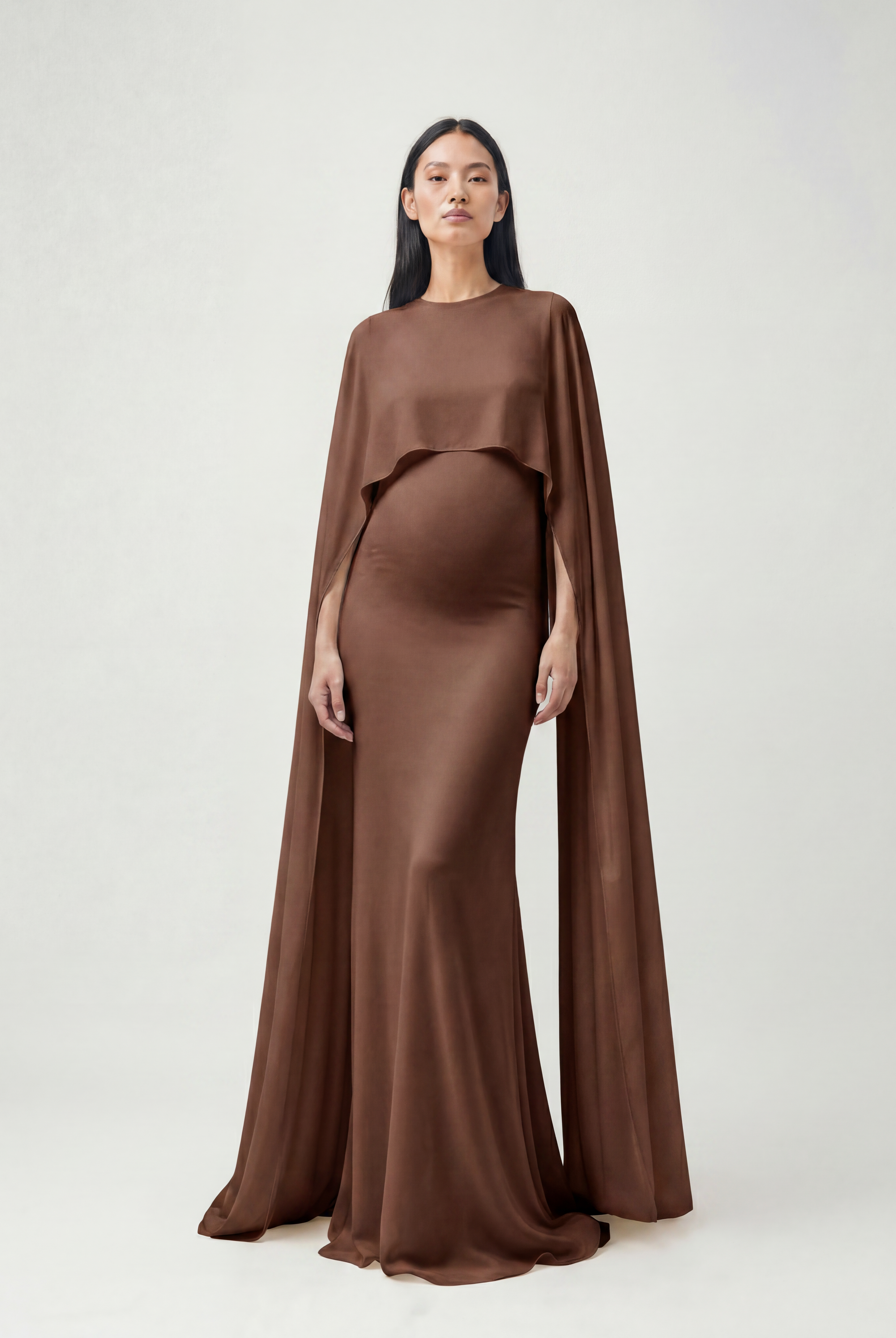 A pregnant model wearing a brown maternity gown with a draped cape overlay, standing against a light gray studio background.
