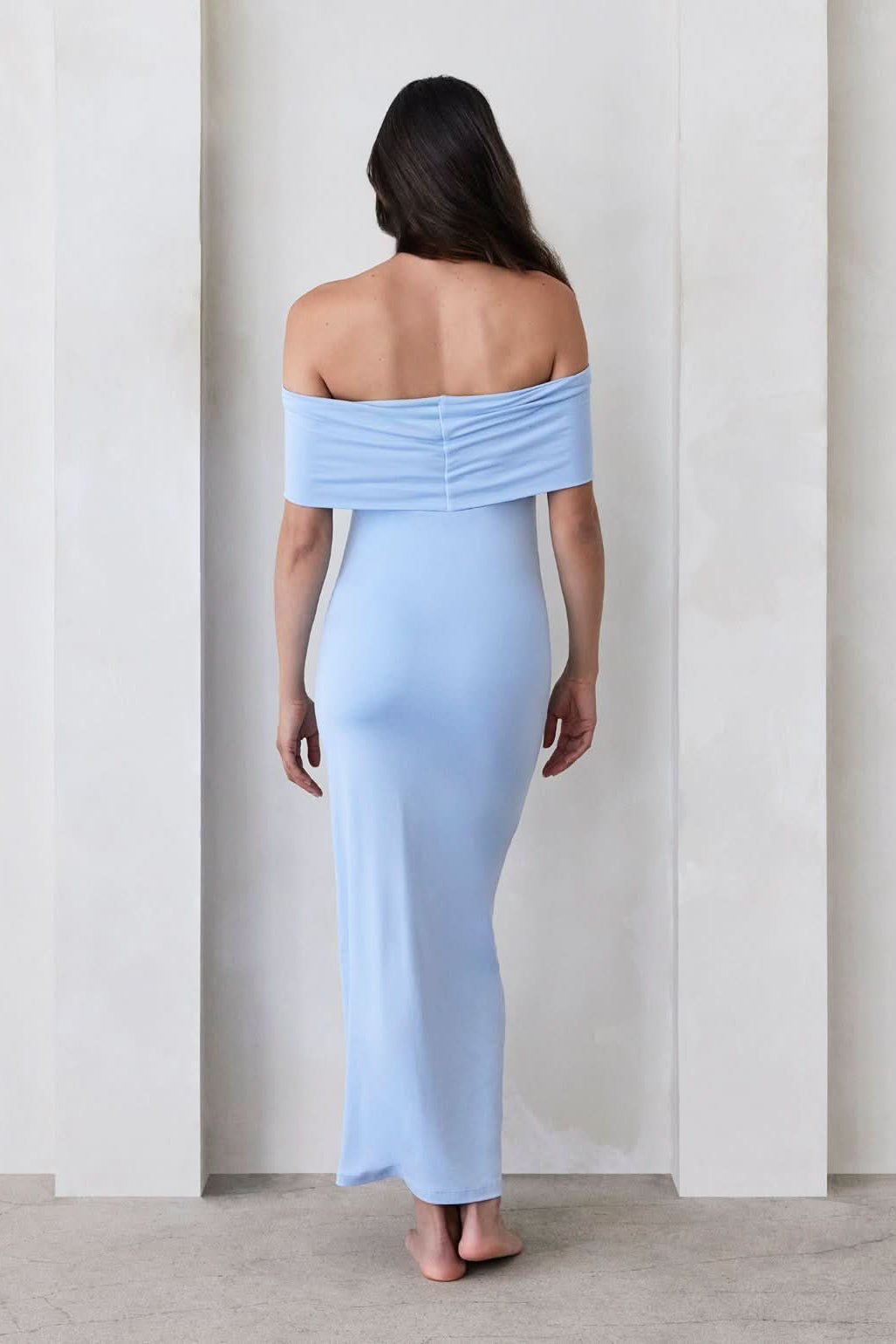 Pregnant woman wearing a Bumpsuit Maternity The Bianca Off Shoulder Maxi Dress in Powder blue standing against a beige wall