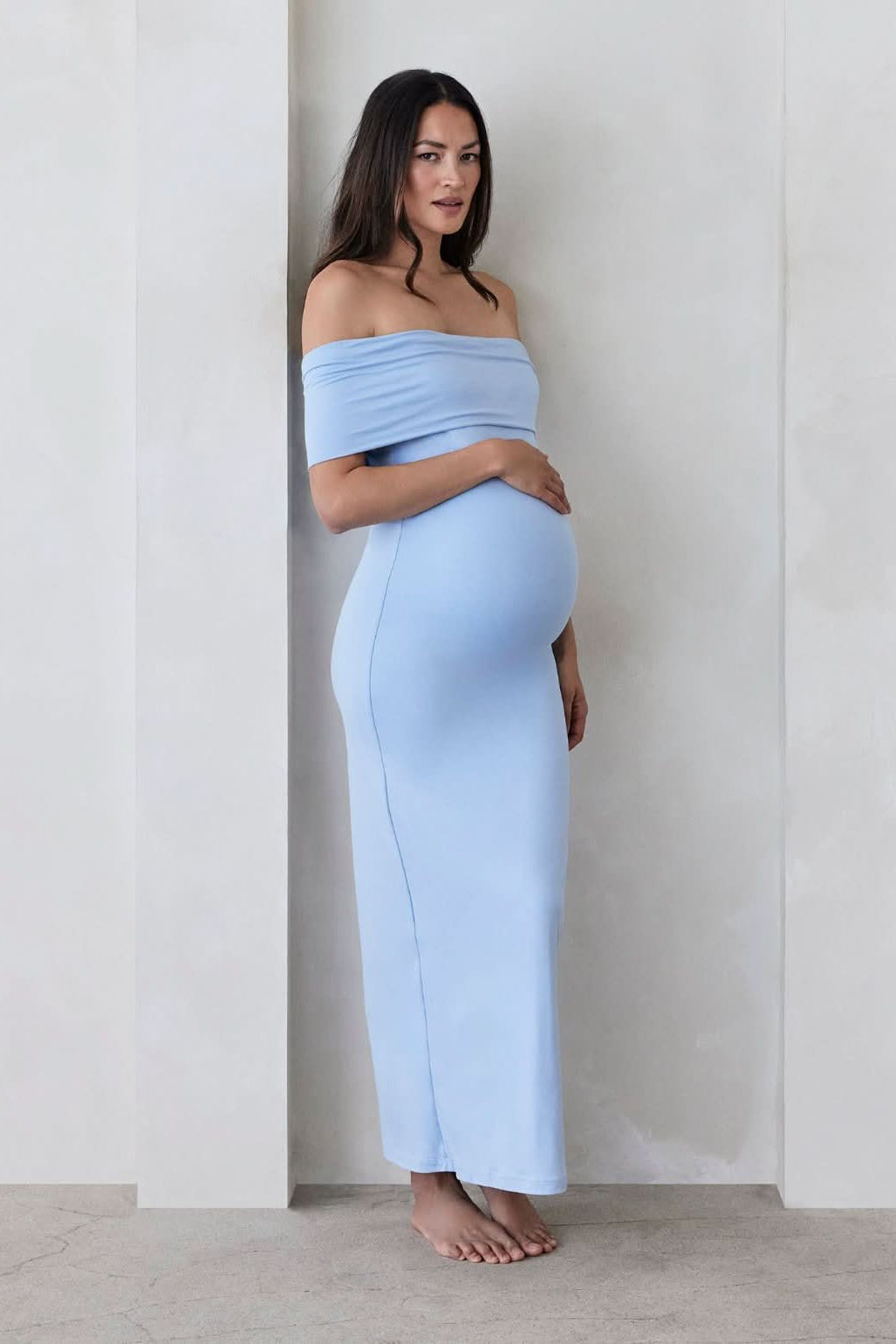 Pregnant woman wearing a Bumpsuit Maternity The Bianca Off Shoulder Maxi Dress in Powder blue standing against a beige wall