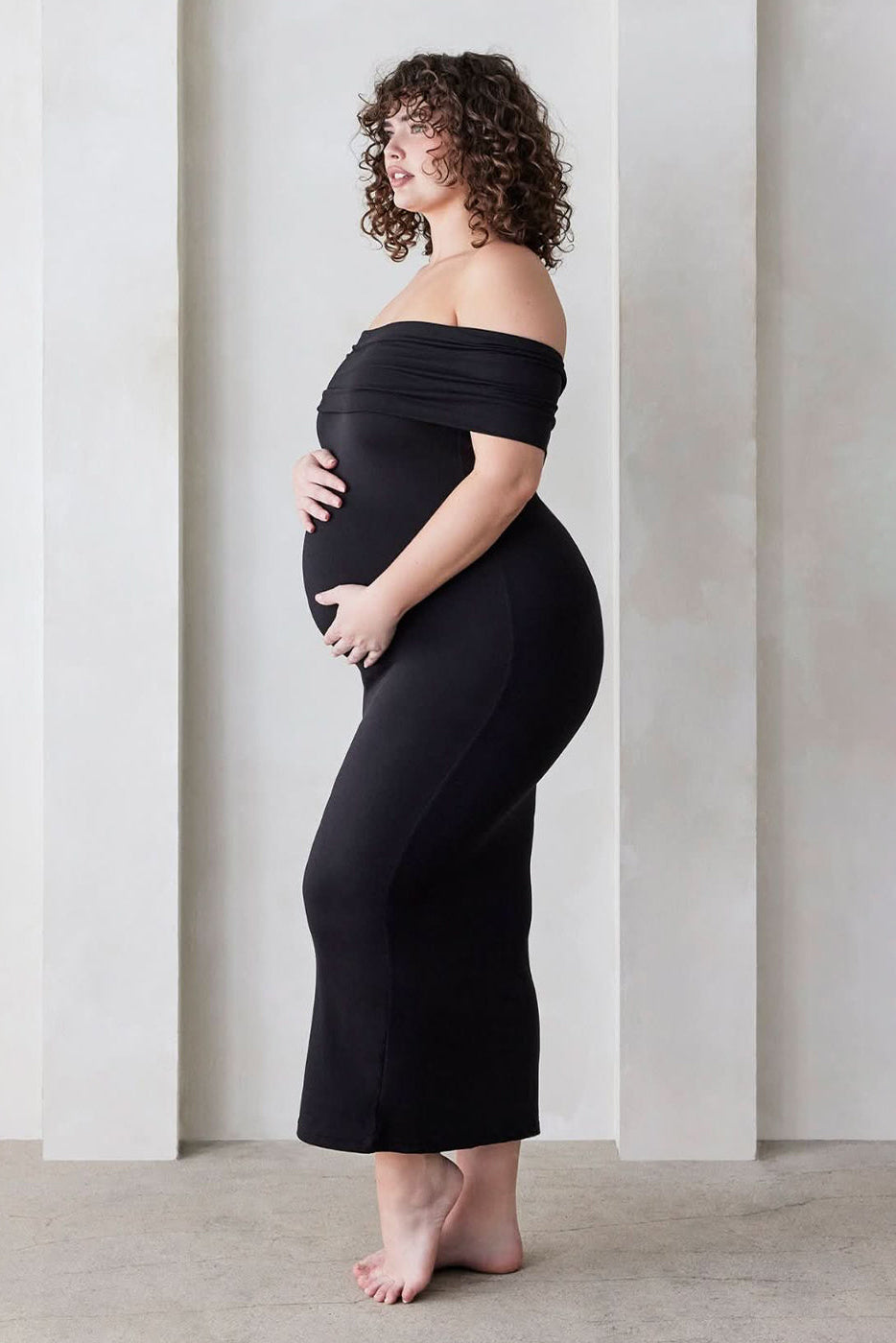 Pregnant woman wearing a Bumpsuit Maternity The Bianca Off Shoulder Maxi Dress in Black standing against a beige wall