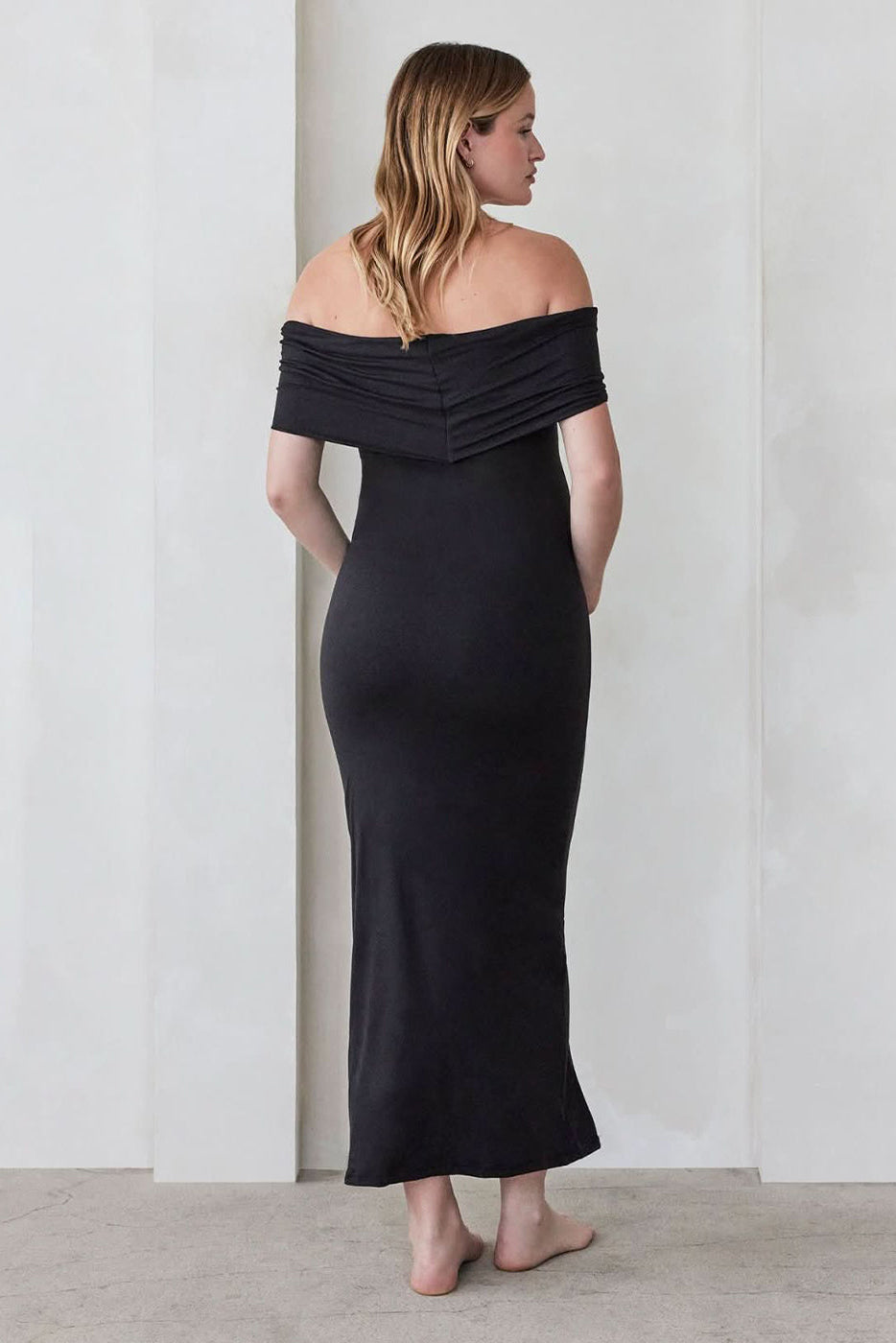 Pregnant woman wearing a Bumpsuit Maternity The Bianca Off Shoulder Maxi Dress in Black standing against a beige wall