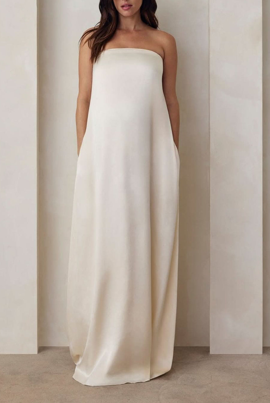 Pregnant model wearing the Bumpsuit brushed satin gown in ivory against a beige feature wall.