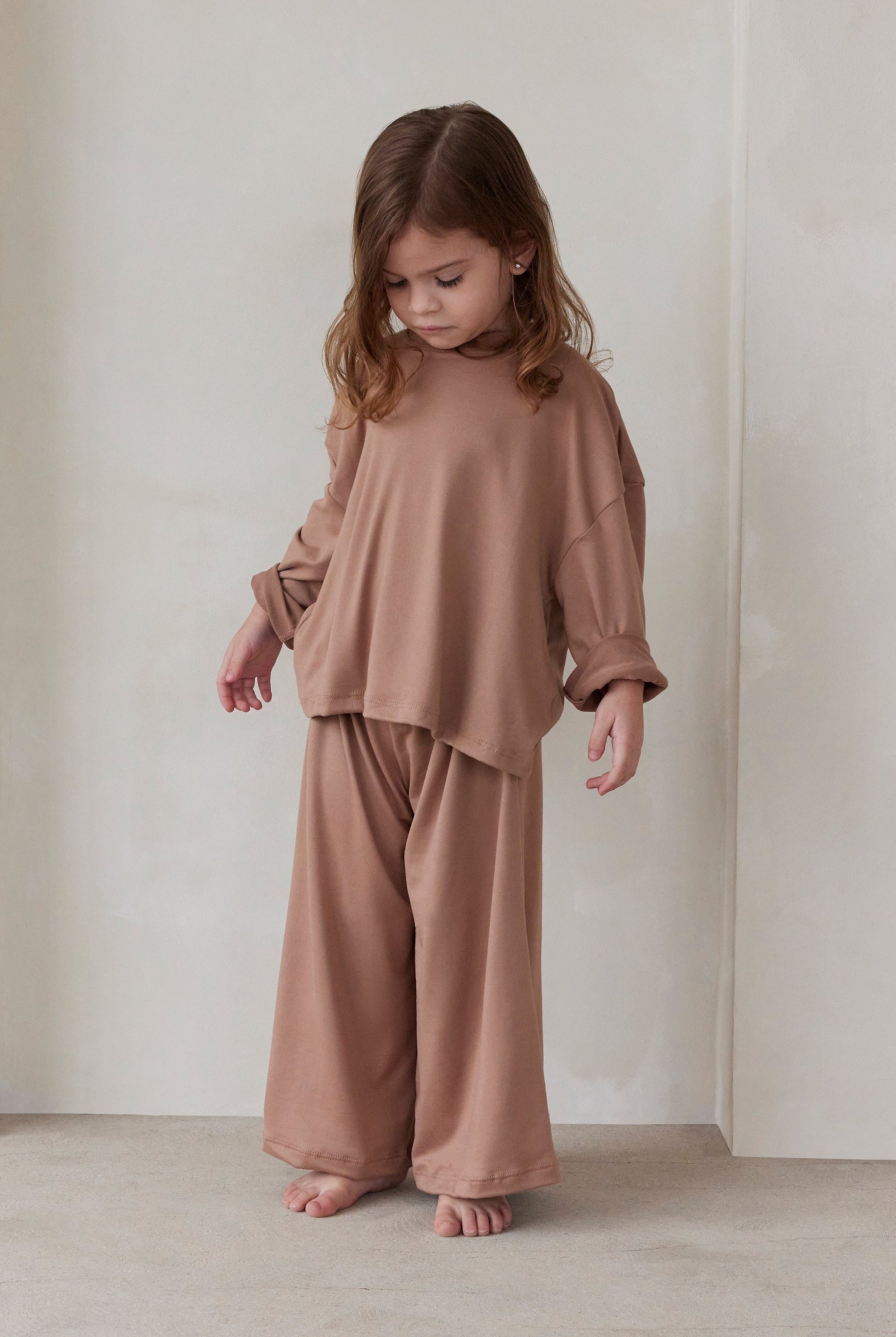 Child wearing a latte brown outfit standing against a plain white wall