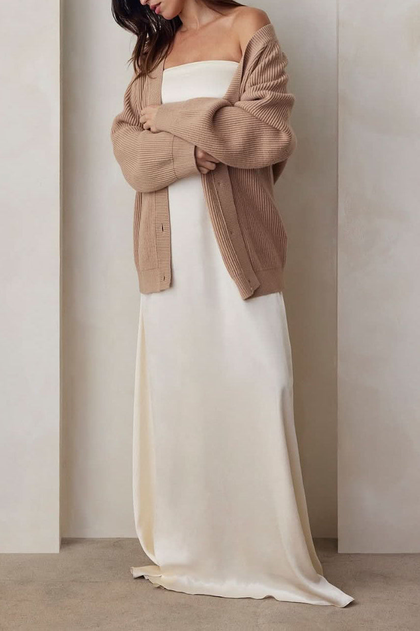 A Pregnant model wearing the Bumpsuit brushed satin gown in ivory and a beige cardigan against a beige feature wall.