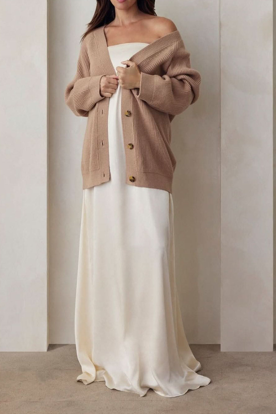 Pregnant model wearing the Bumpsuit brushed satin gown in ivory and a beige knit oversized cardigan against a beige feature wall.