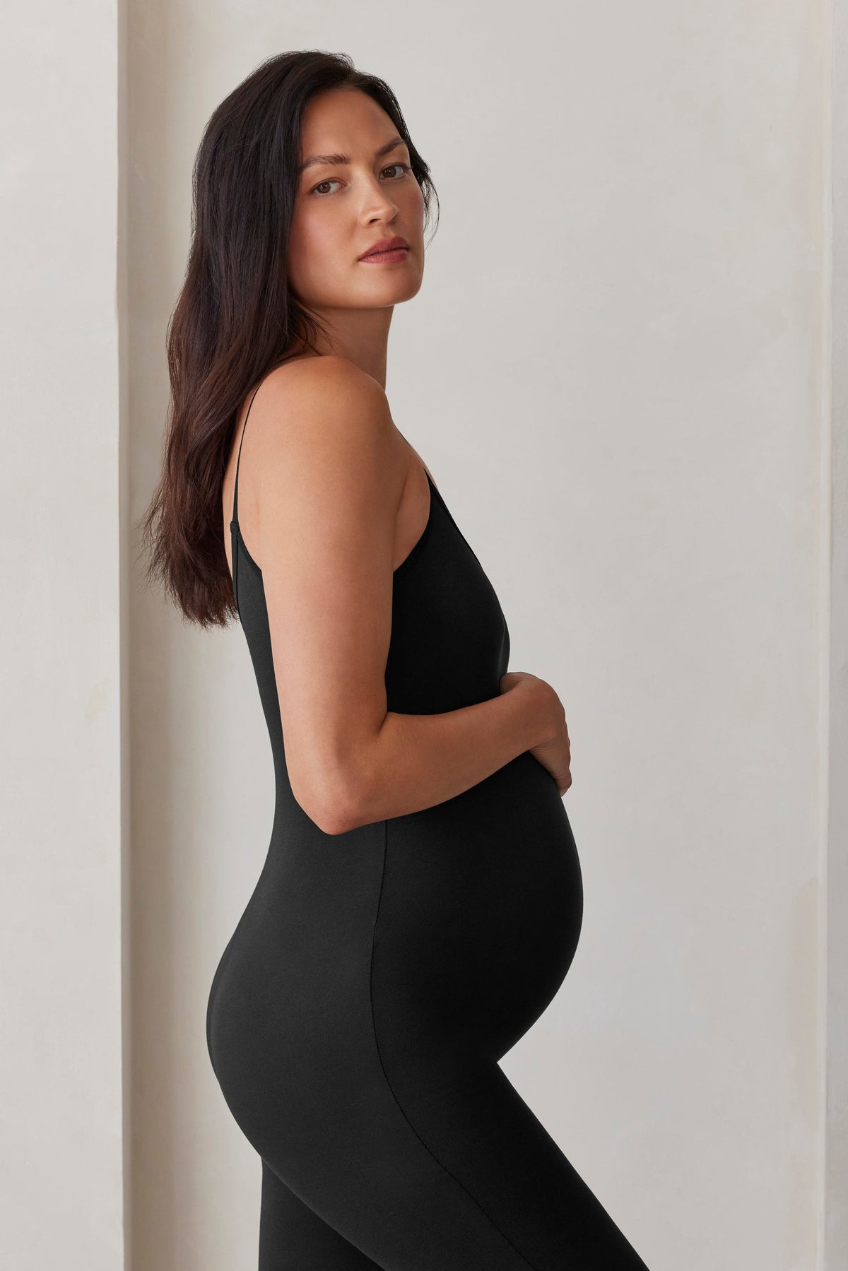 Pregnant woman wearing the Bumpsuit Kate Maternity Jumpsuit in Black against against a beige feature wall