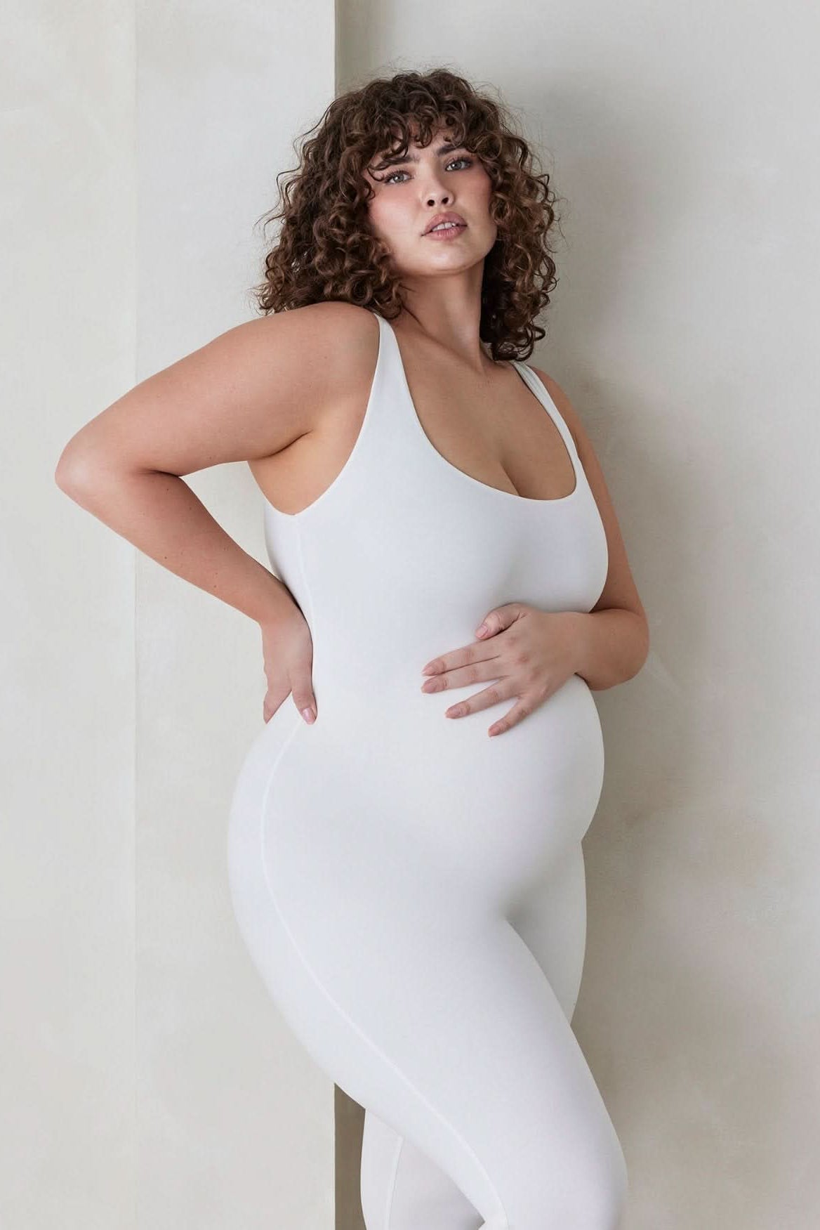 Pregnant woman wearing the Lucy maternity scoop neck unitard in Ivory by Bumpsuit set against a feature wall