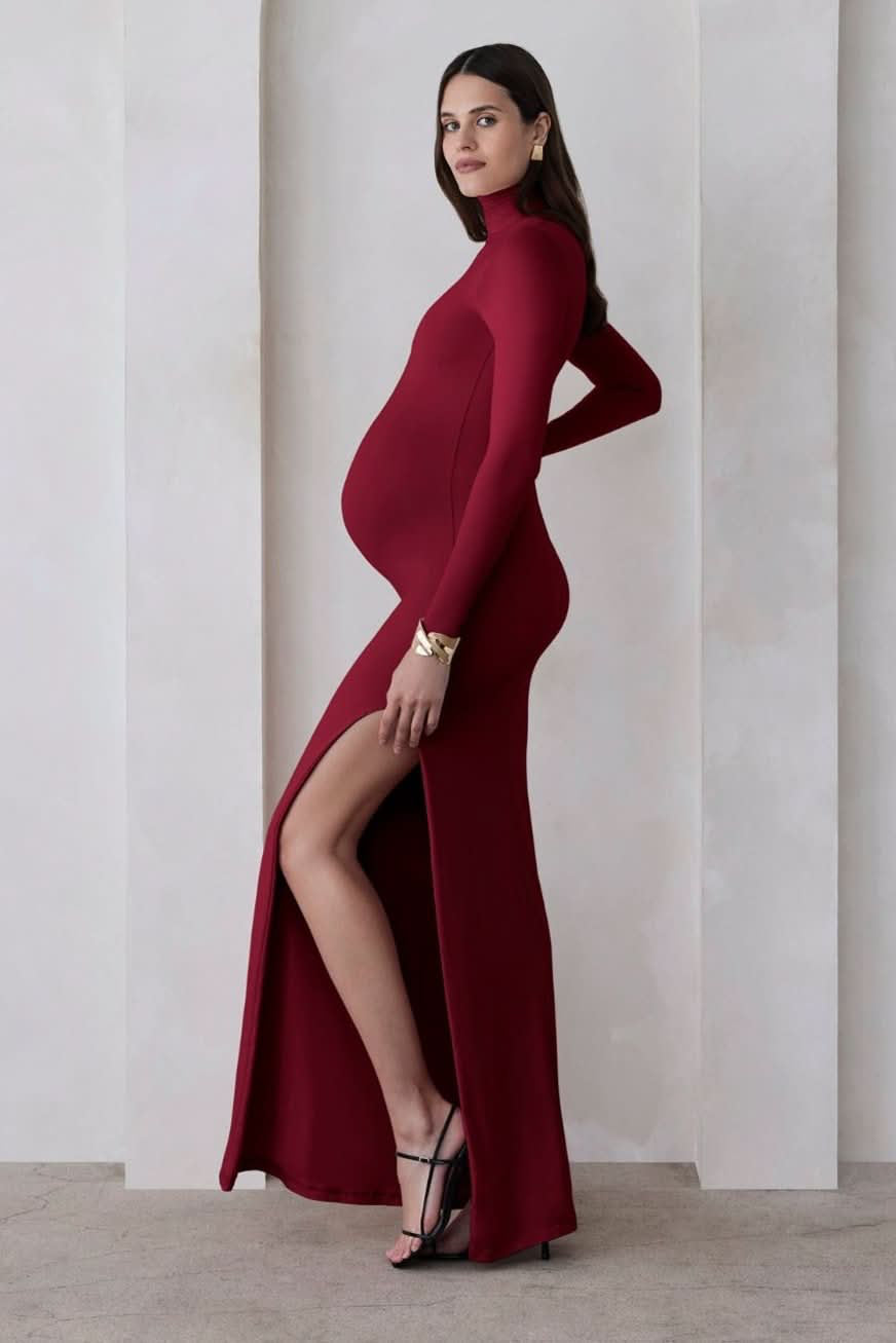 A pregnant woman wearing a burgundy turtleneck maternity maxi dress with long sleeves and a thigh-high slit on one side standing against a beige feature wall