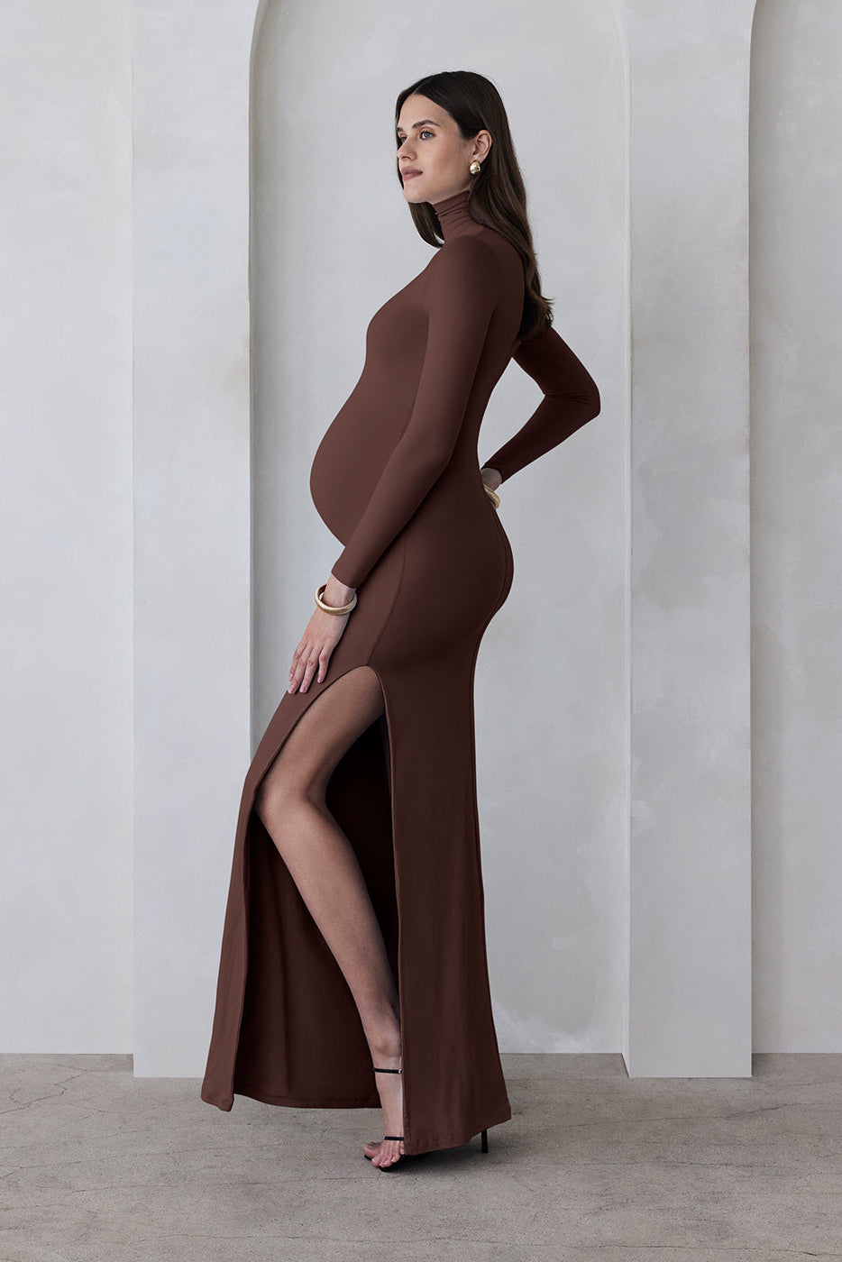 A pregnant woman wearing a chocolate brown turtleneck maternity maxi dress with long sleeves and a thigh-high slit on one side standing against a beige feature wall