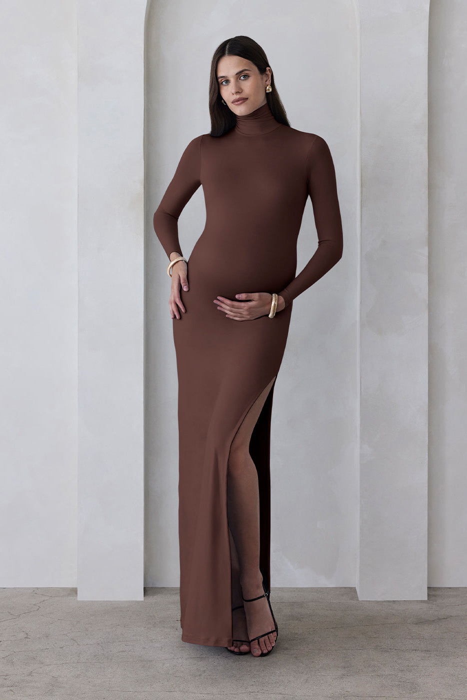 A pregnant woman wearing a chocolate brown turtleneck maternity maxi dress with long sleeves and a thigh-high slit on one side standing against a beige feature wall