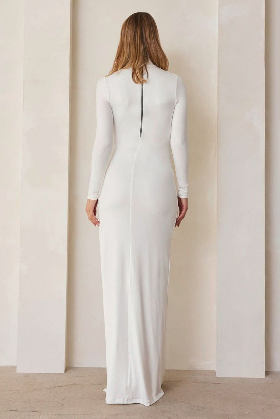 Back view of The Monica Maternity Dress in ivory showcasing long sleeves, turtleneck and center-back zipper against beige wall