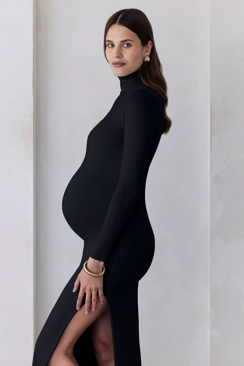 A pregnant woman wearing a black turtleneck maternity maxi dress with long sleeves and a thigh-high slit on one side standing against a feature wall