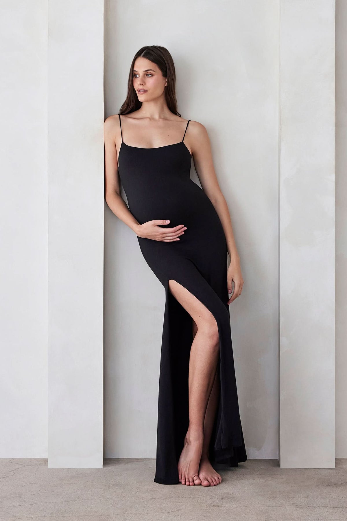 Pregnant woman wearing The Rosie maternity Square Neck Sleeveless Maxi Dress with side slit in black by Bumpsuit, standing beige against a feature wall