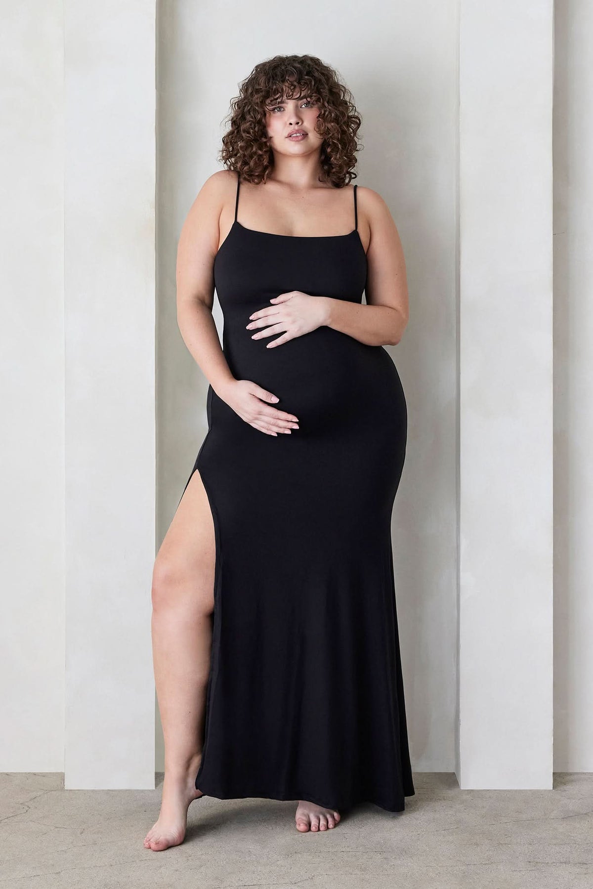 Pregnant woman wearing The Rosie maternity Square Neck Sleeveless Maxi Dress with side slit in black by Bumpsuit, standing beige against a feature wall