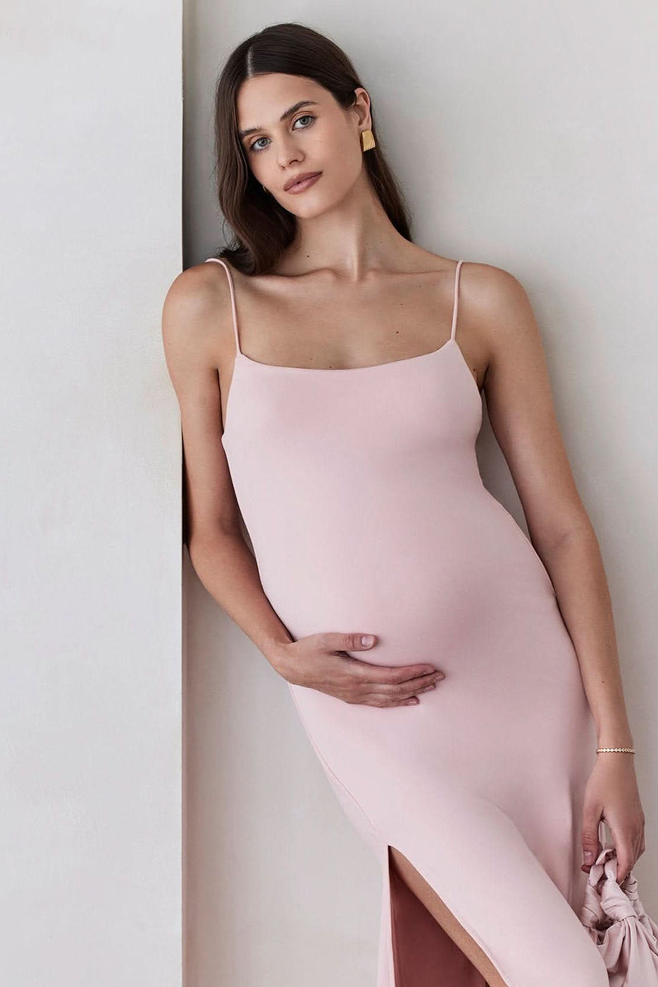 Pregnant woman wearing The Rosie maternity Square Neck Sleeveless Maxi Dress with side slit in dusty pink by Bumpsuit, holding a bag standing beige against a feature wall