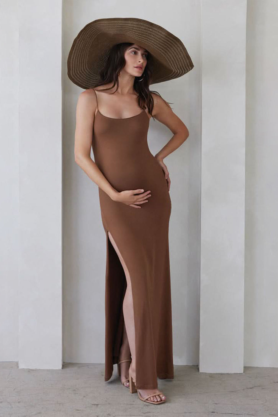 Pregnant woman wearing The Rosie maternity Square Neck Sleeveless Maxi Dress with side slit in Mocha brown by Bumpsuit, wearing a hat standing beige against a feature wall
