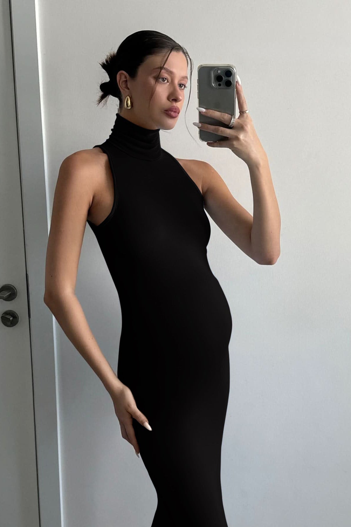 Pregnant woman wears the Serena sleeveless turtleneck maxi dress by Bumpsuit in black, holding a phone