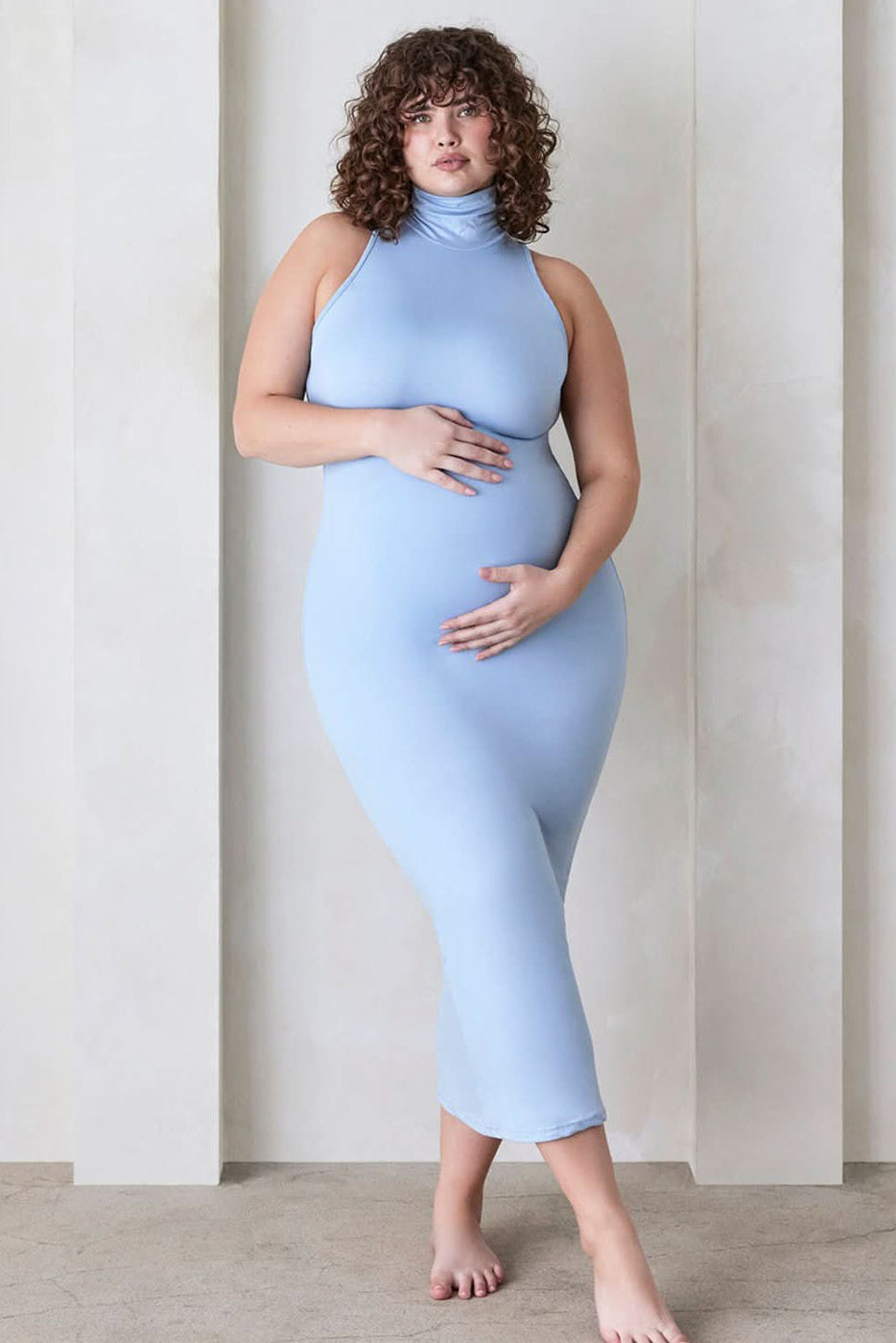 Pregnant woman wears the Serena sleeveless turtleneck maxi dress by Bumpsuit in powder blue, standing against a beige feature wall