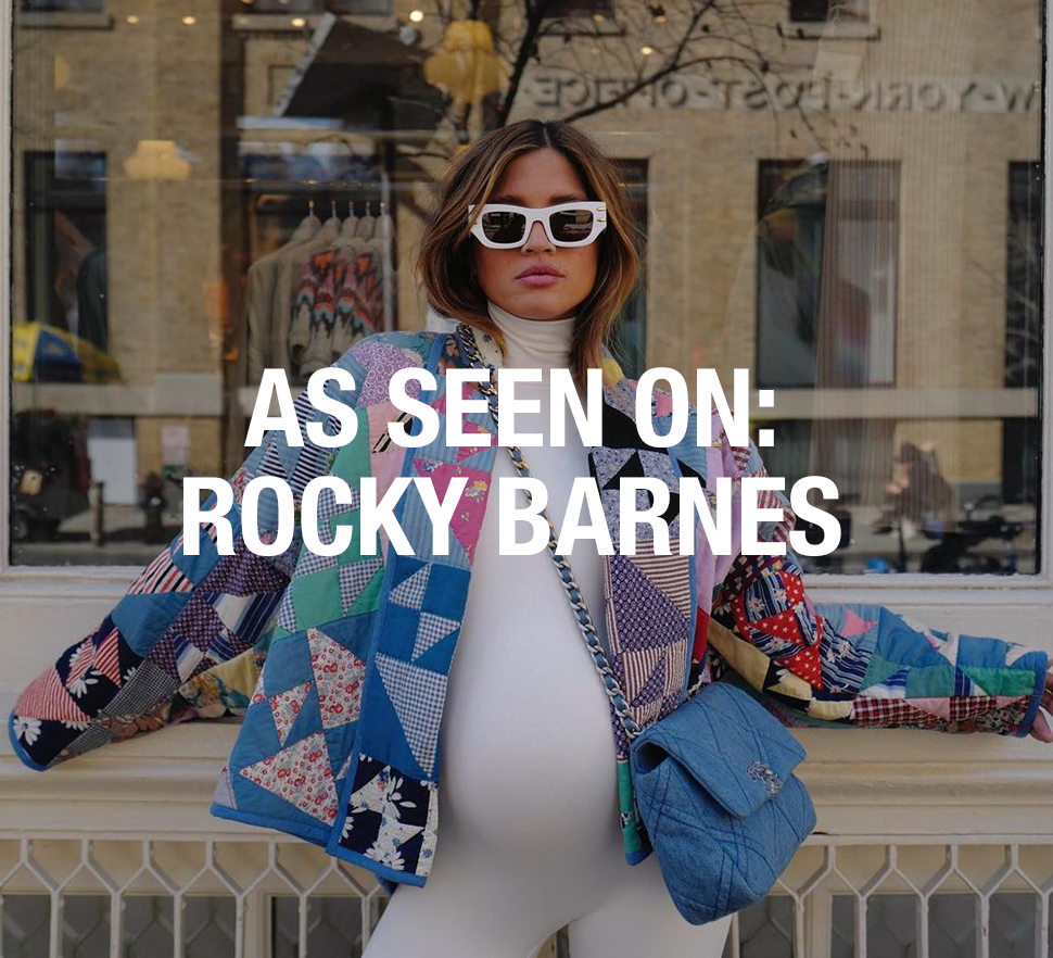 Rocky Barnes poses in a white maternity bodysuit, patchwork jacket, sunglasses, and blue bag outside a storefront