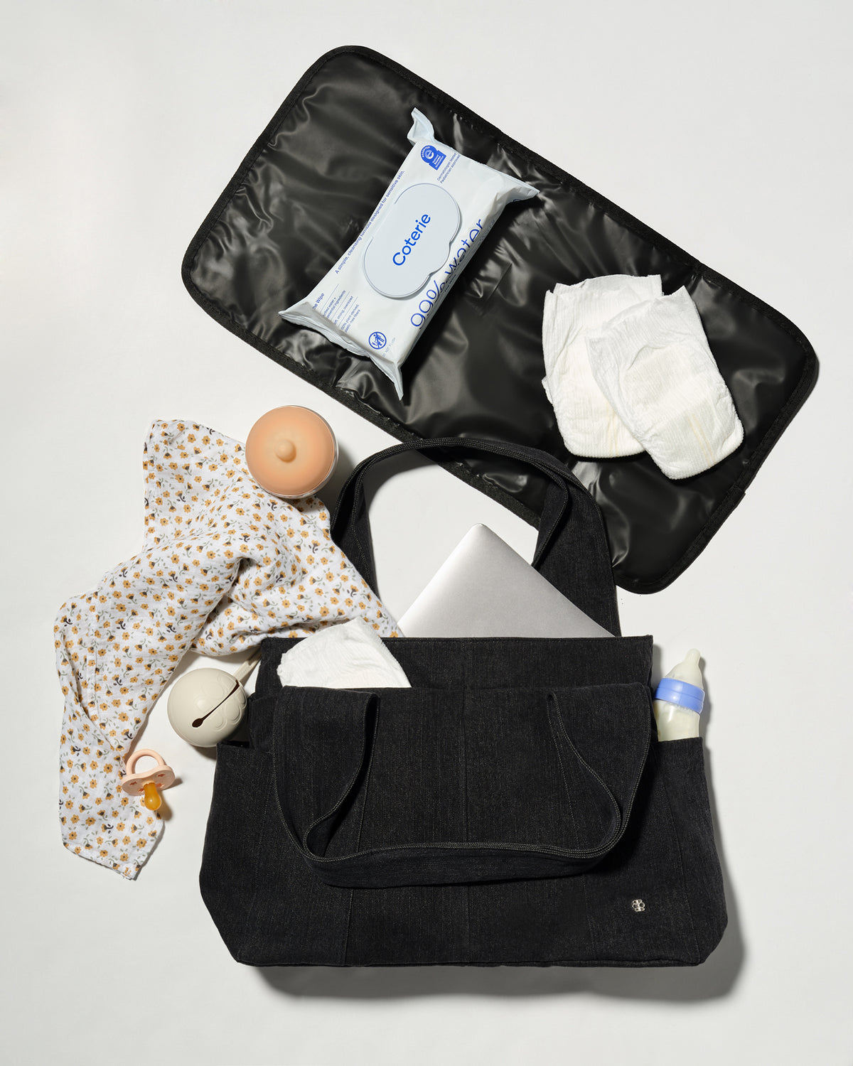 A black diaper bag shown open in a flat-lay arrangement, filled with baby essentials including a patterned baby shirt, wipes, diapers, a pacifier, and a bottle, on a light grey background.