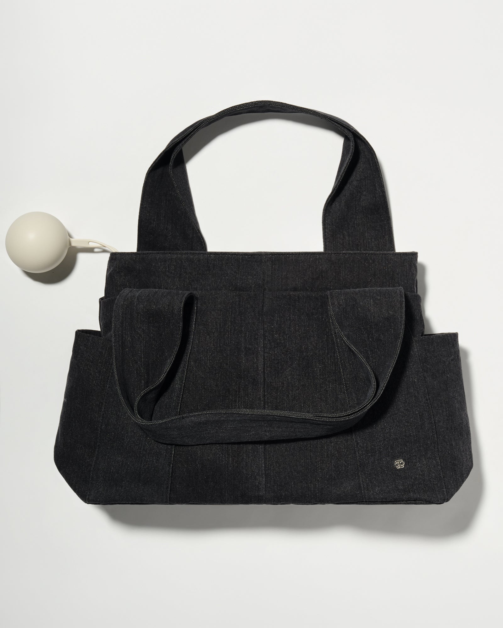 A black denim fabric diaper bag with dual top handles and front slip pockets, shown on a plain light background with a small pacifier holder on the left.