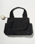 A black denim fabric diaper bag with dual top handles and front slip pockets, shown on a plain light background with a small pacifier holder on the left.