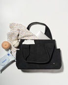 A black denim fabric diaper bag laid flat on a light grey background, styled with a patterned scarf, a white laptop, a baby bottle, a pacifier holder, and a packet of baby wipes.