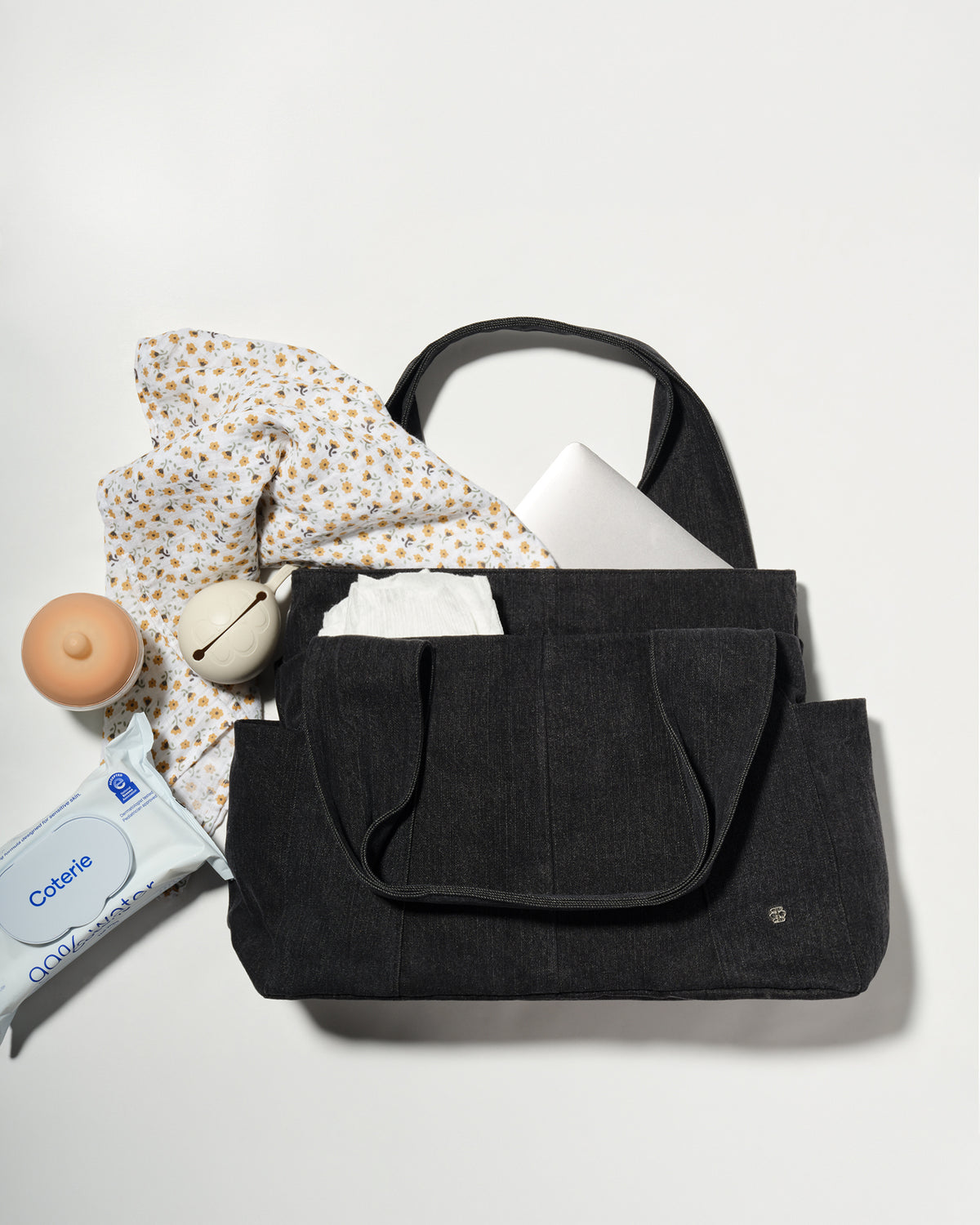 A black denim fabric diaper bag laid flat on a light grey background, styled with a patterned scarf, a white laptop, a baby bottle, a pacifier holder, and a packet of baby wipes.