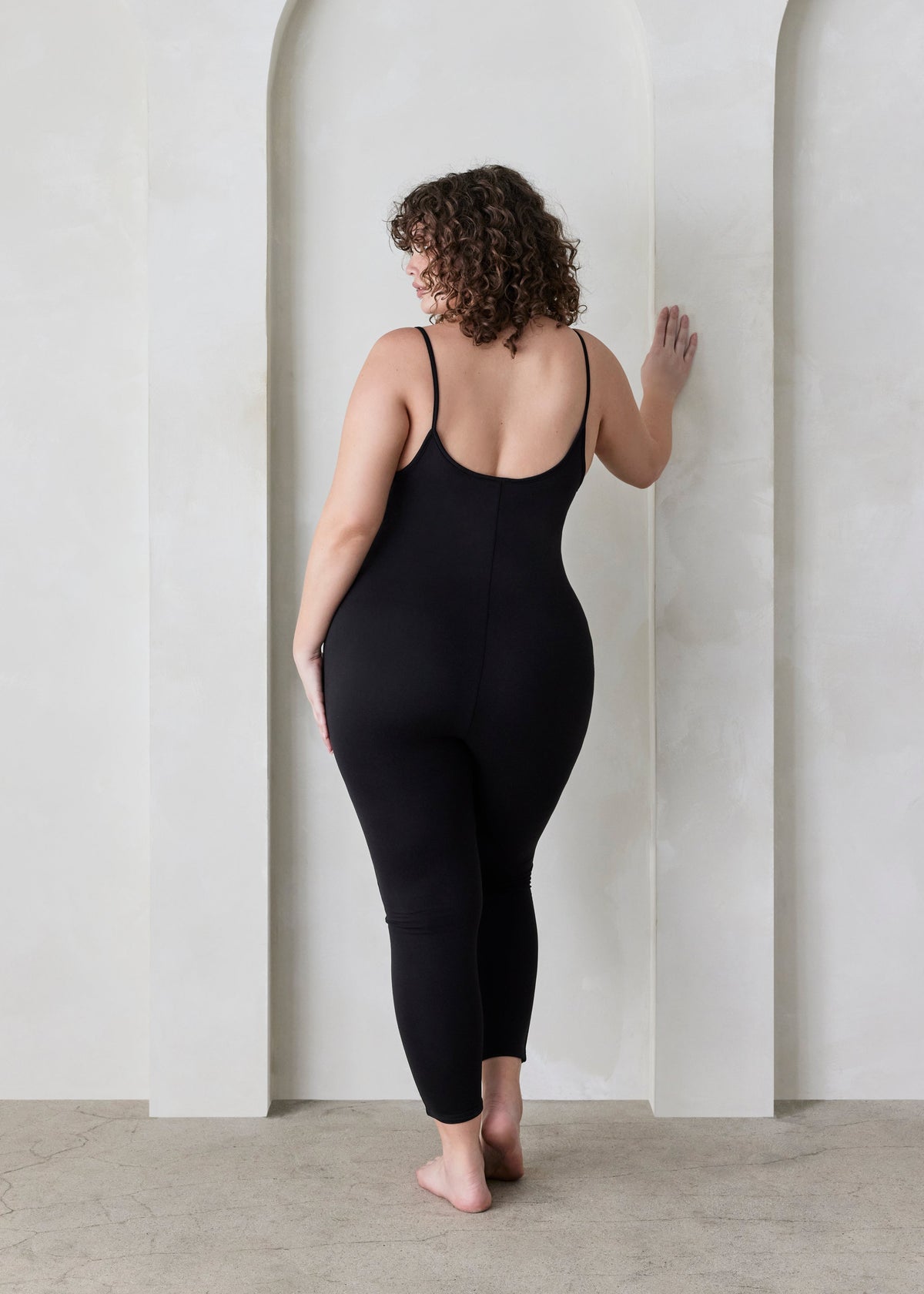 Back view of a woman wearing a Bumpsuit black sleeveless jumpsuit called the Kate  standing in a minimalistic setting.