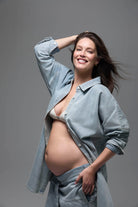 Pregnant woman smiling wearing Bumpsuits light blue denim button up shirt and jeans against a gray background