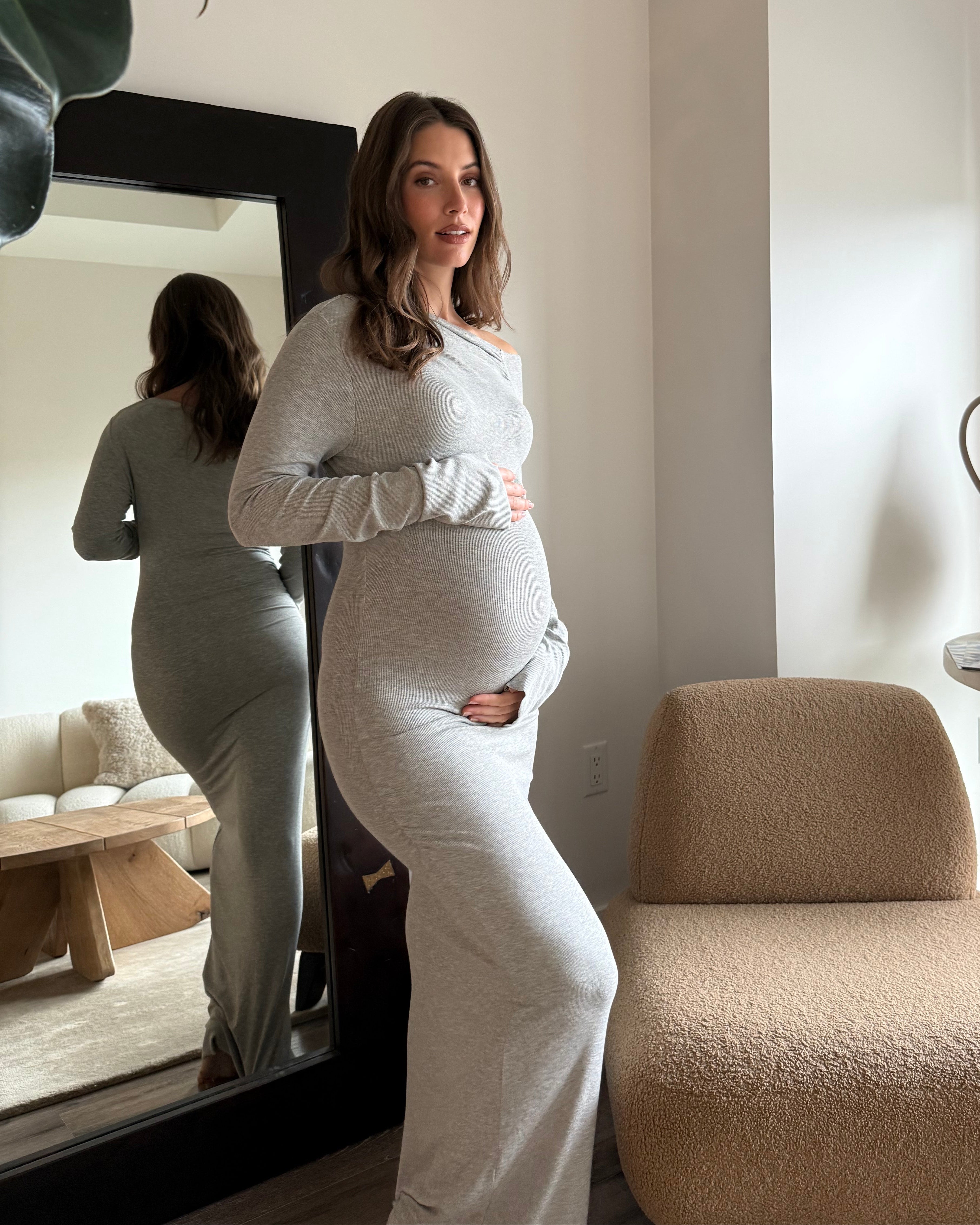 A pregnant woman wearing a heather gray  Bumpsuit soft rib long-sleeve maternity dress stands in a modern room, posing in front of a large mirror that reflects her back. The room features a beige boucle chair, a side table with ceramic vases, and a large green plant in the foreground.