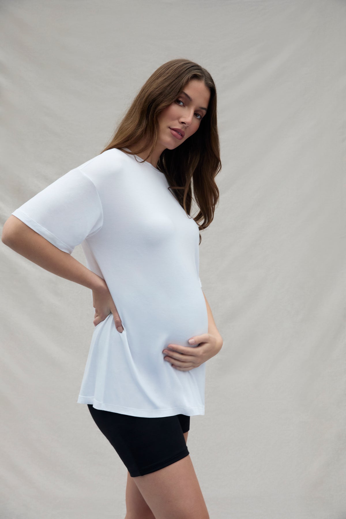 Pregnant woman wearing a white top and black shorts against a plain background
