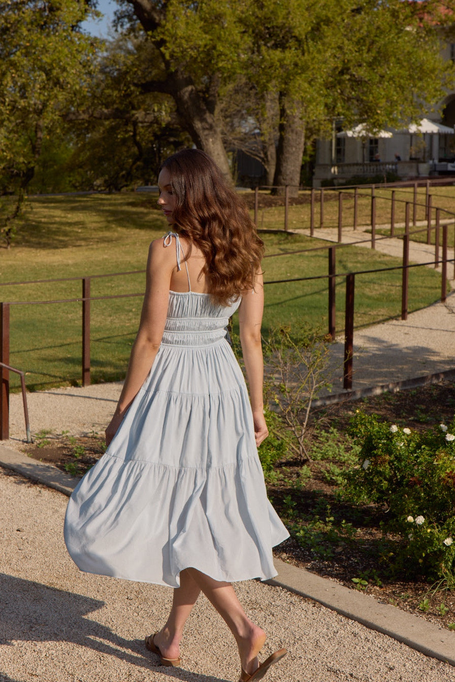 Back view of ice blue shirred midi poplin maternity dress showing a fitted bodice and tiered skirt in a garden