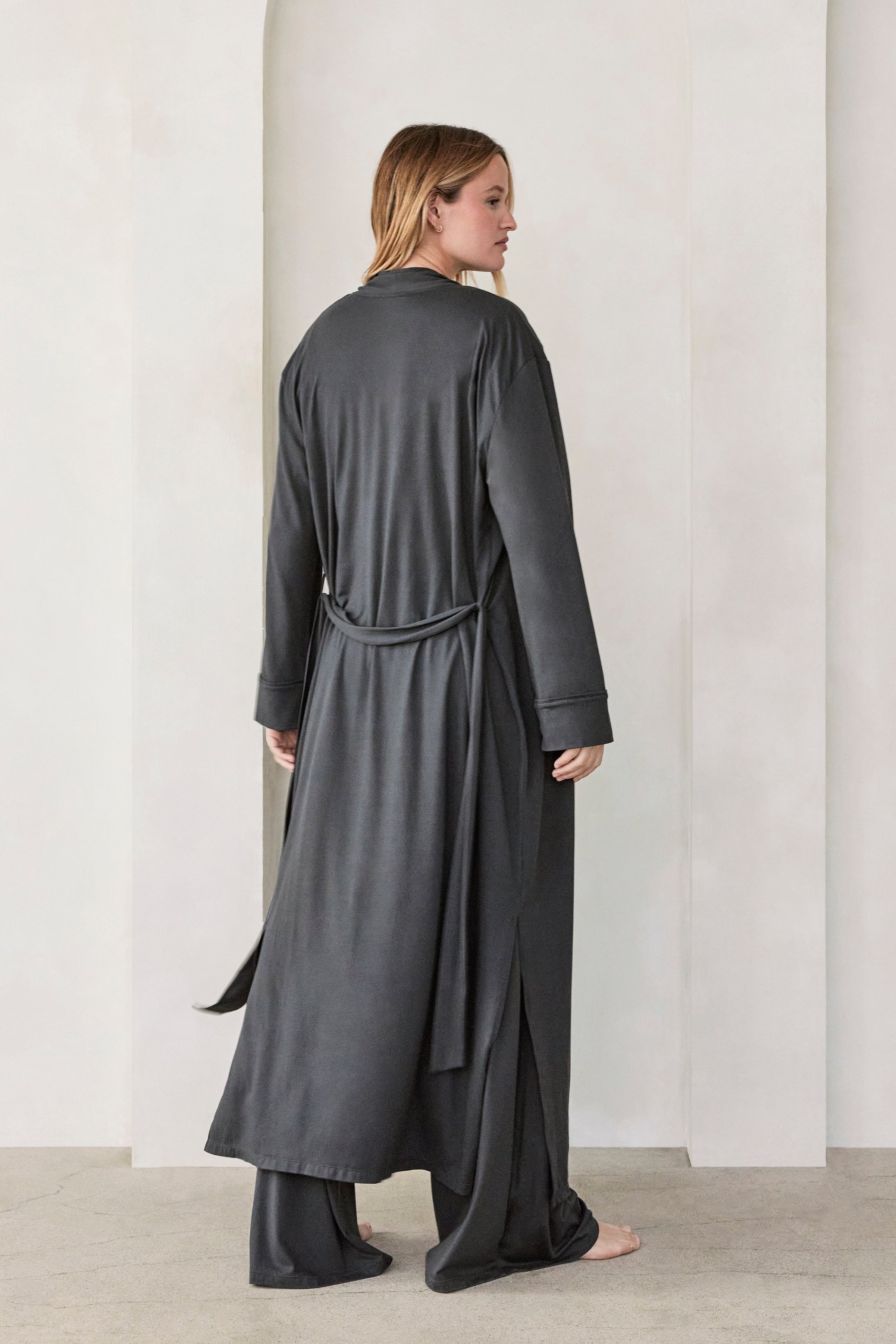 Woman wearing a dark grey slate Bumpsuit Cloud outfit standing in a minimalistic room with beige walls and arches.