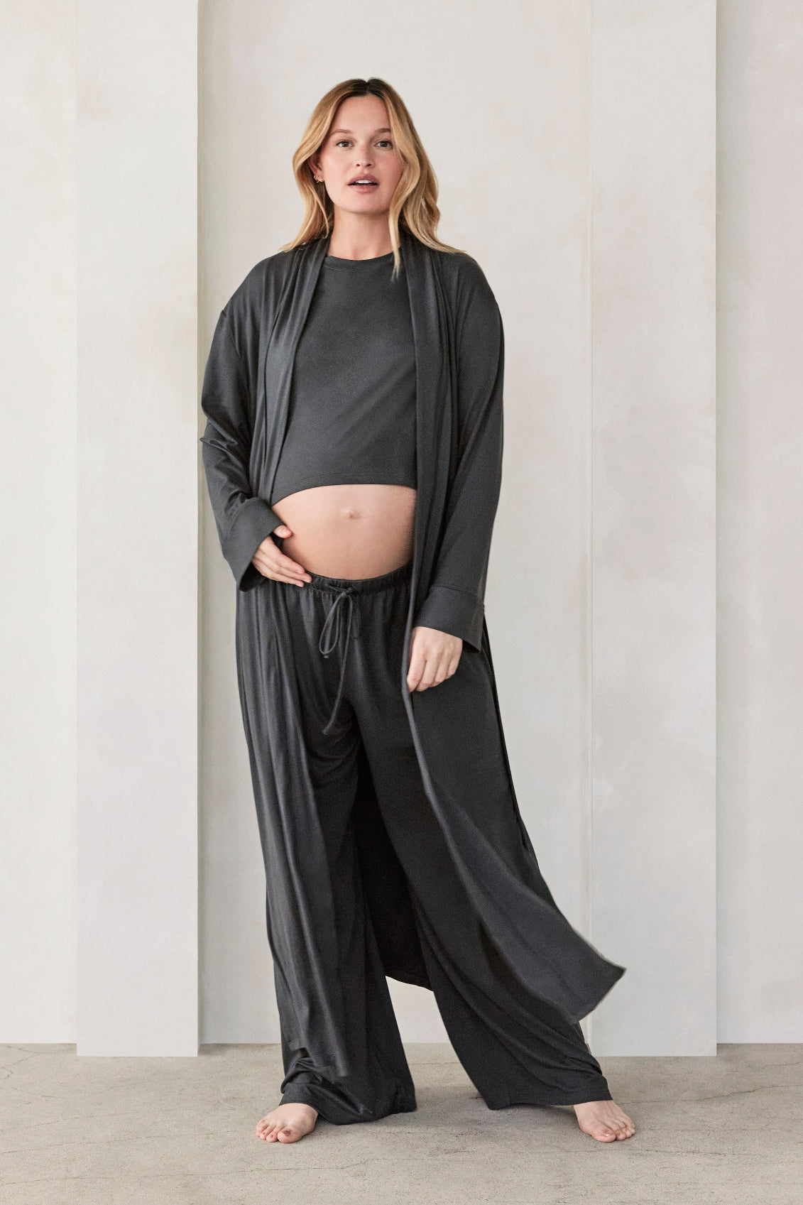 Woman wearing a dark grey slate Bumpsuit Cloud outfit standing in a minimalistic room with beige walls and arches.
