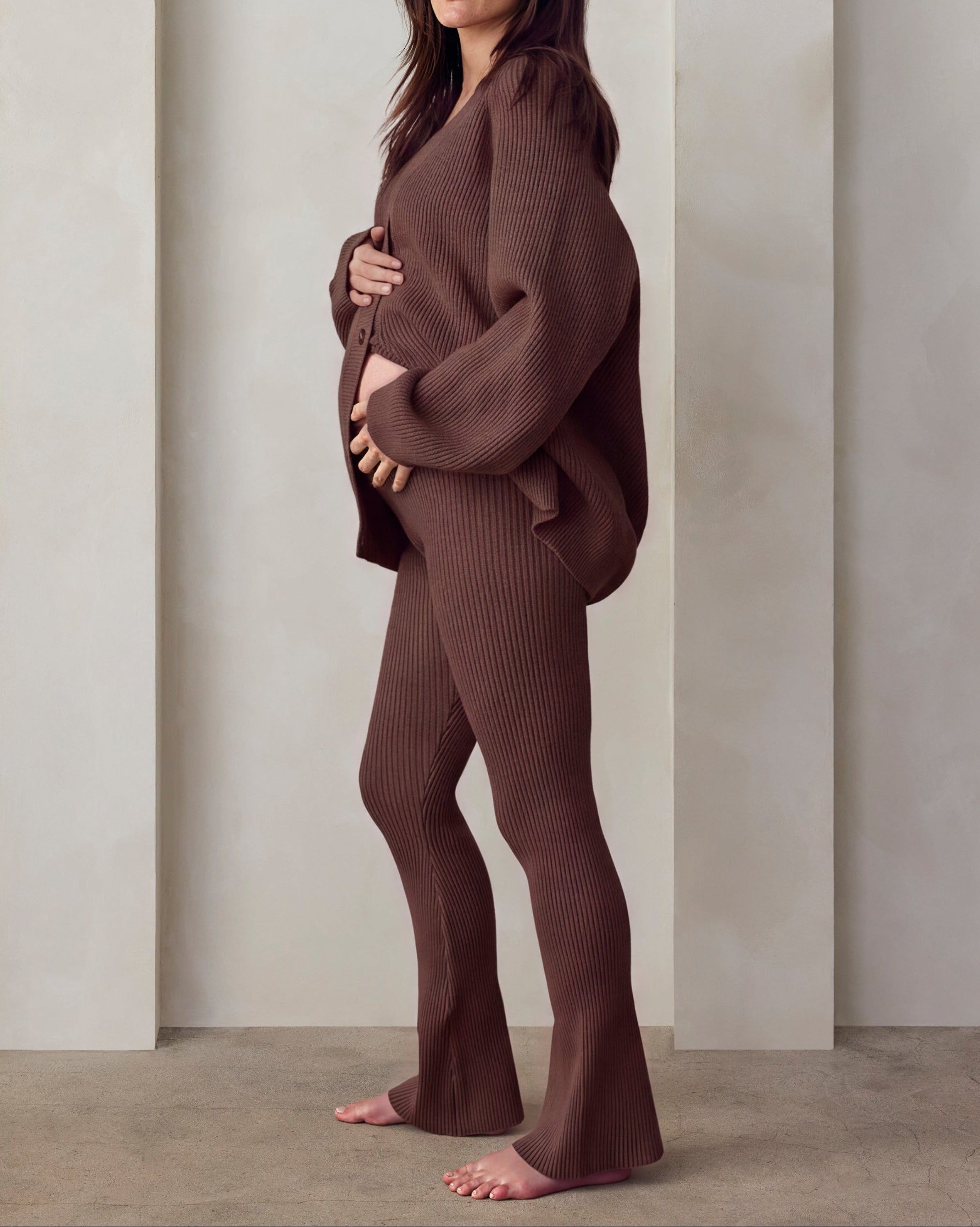 A woman stands barefoot in a minimalist studio setting with three arched recesses on a cream-colored wall. She is wearing a matching ribbed brown lounge set cyle top and flared pants.