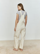Back view of a woman wearing beige overalls against a plain background