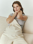Pregnant woman sitting on a beige couch wearing a Bumpsuit baby mama tee and beige overalls.
