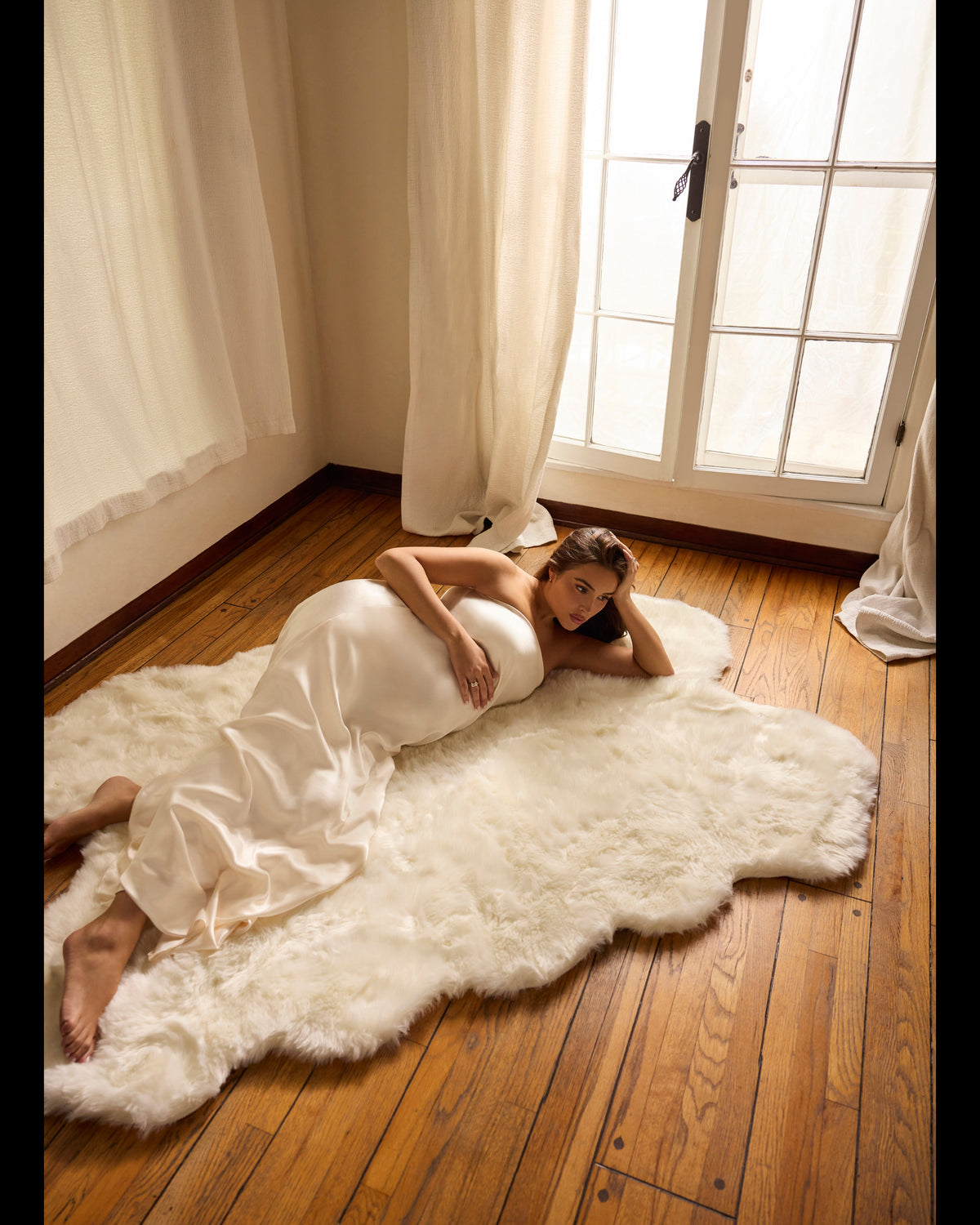 A woman in a white satin gown reclines on a fluffy white sheepskin rug on a hardwood floor, positioned near a window with cream curtains.