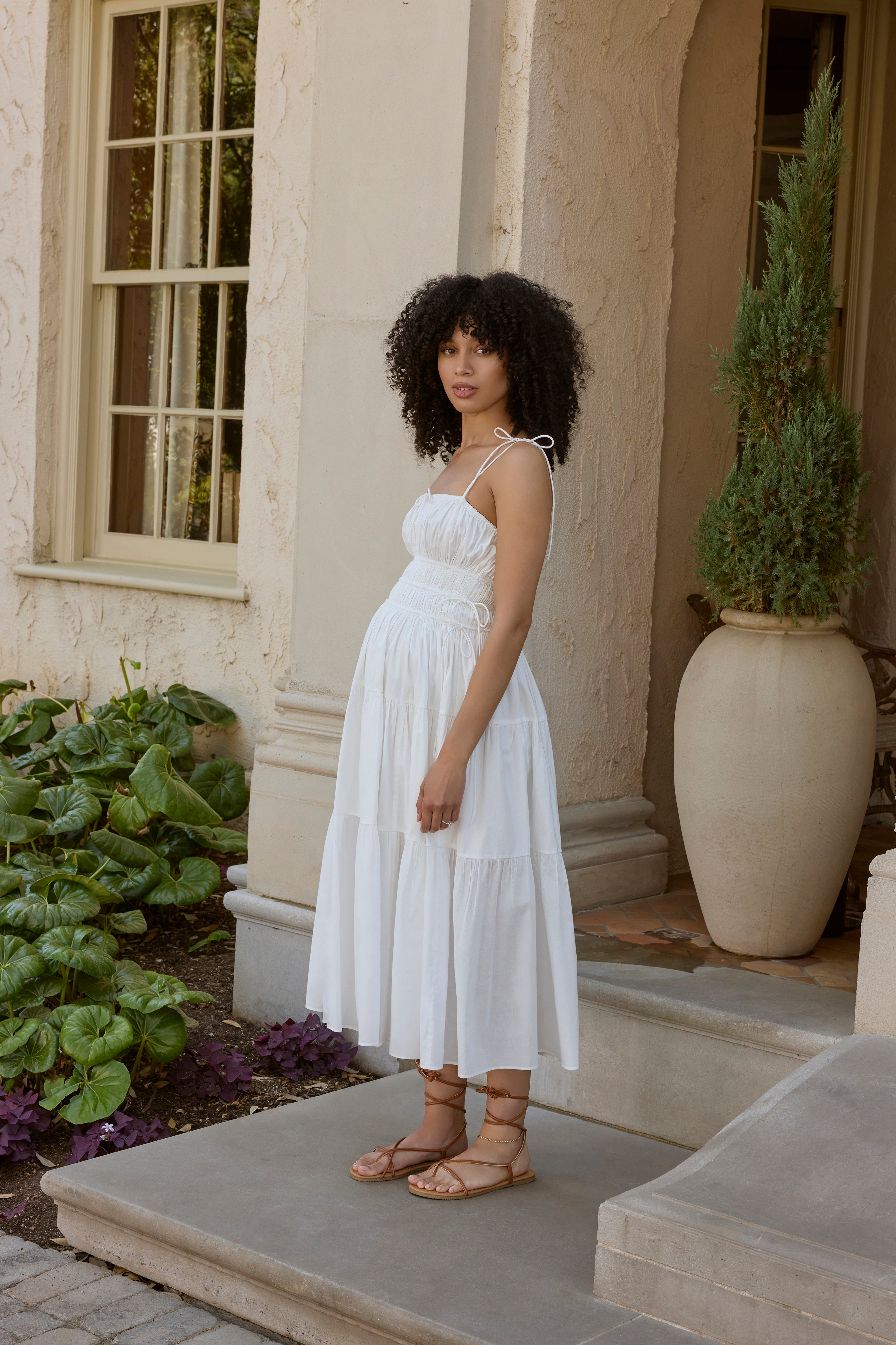 Woman in a white dress standing on a patio with plants and a building in the background