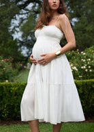 Pregnant woman in a white dress standing outdoors with greenery in the background